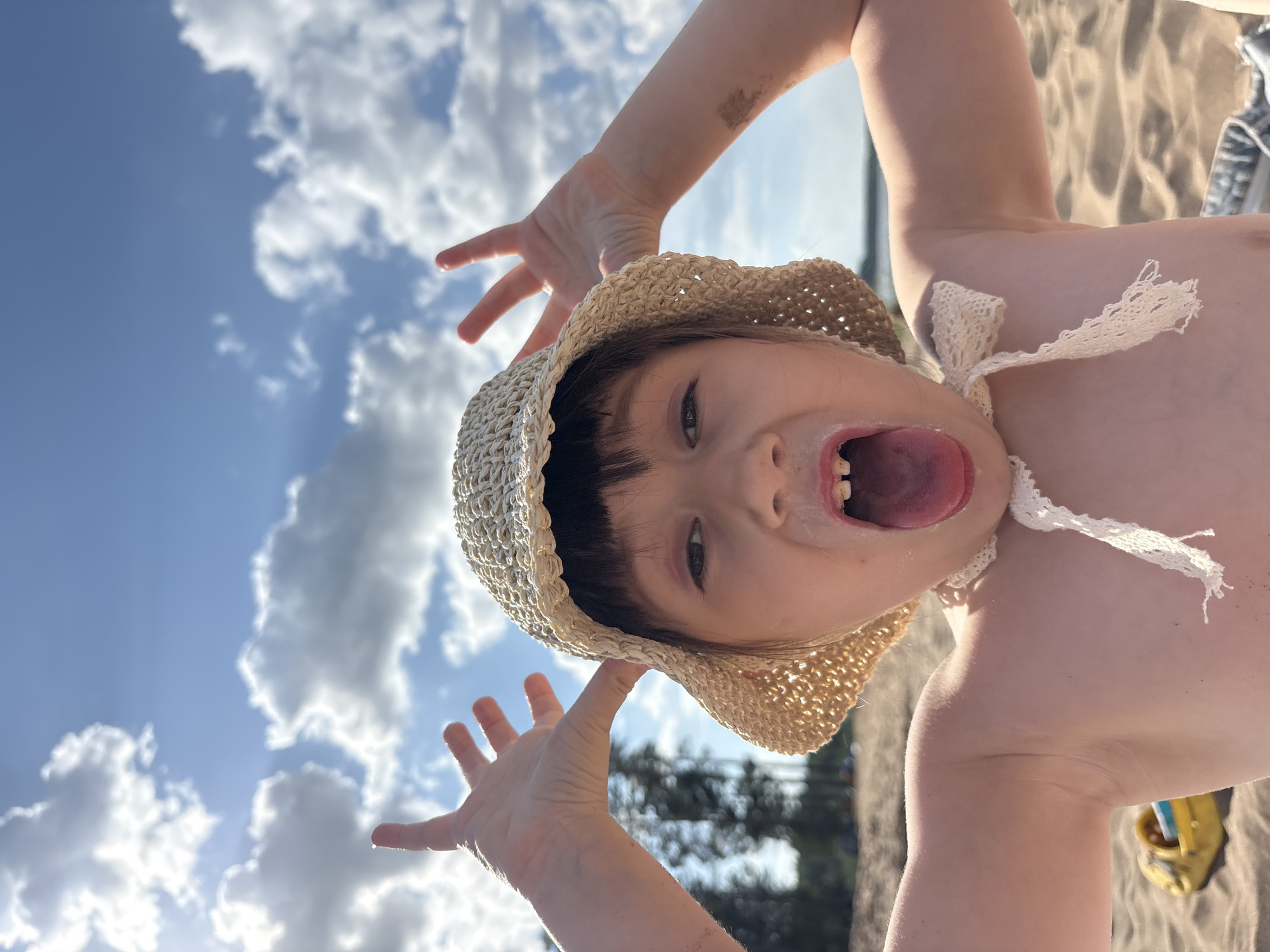 A child lies on a sandy surface, possibly a beach, with a blue sky and clouds visible in the background. The child wears a straw sun hat with a white lace tie. The child has their arms raised, with hands open and fingers spread. The child's mouth is open, tongue extended. The background also includes foliage and possibly a glimpse of a shoe. The location is near Chaikovsky, Russia.