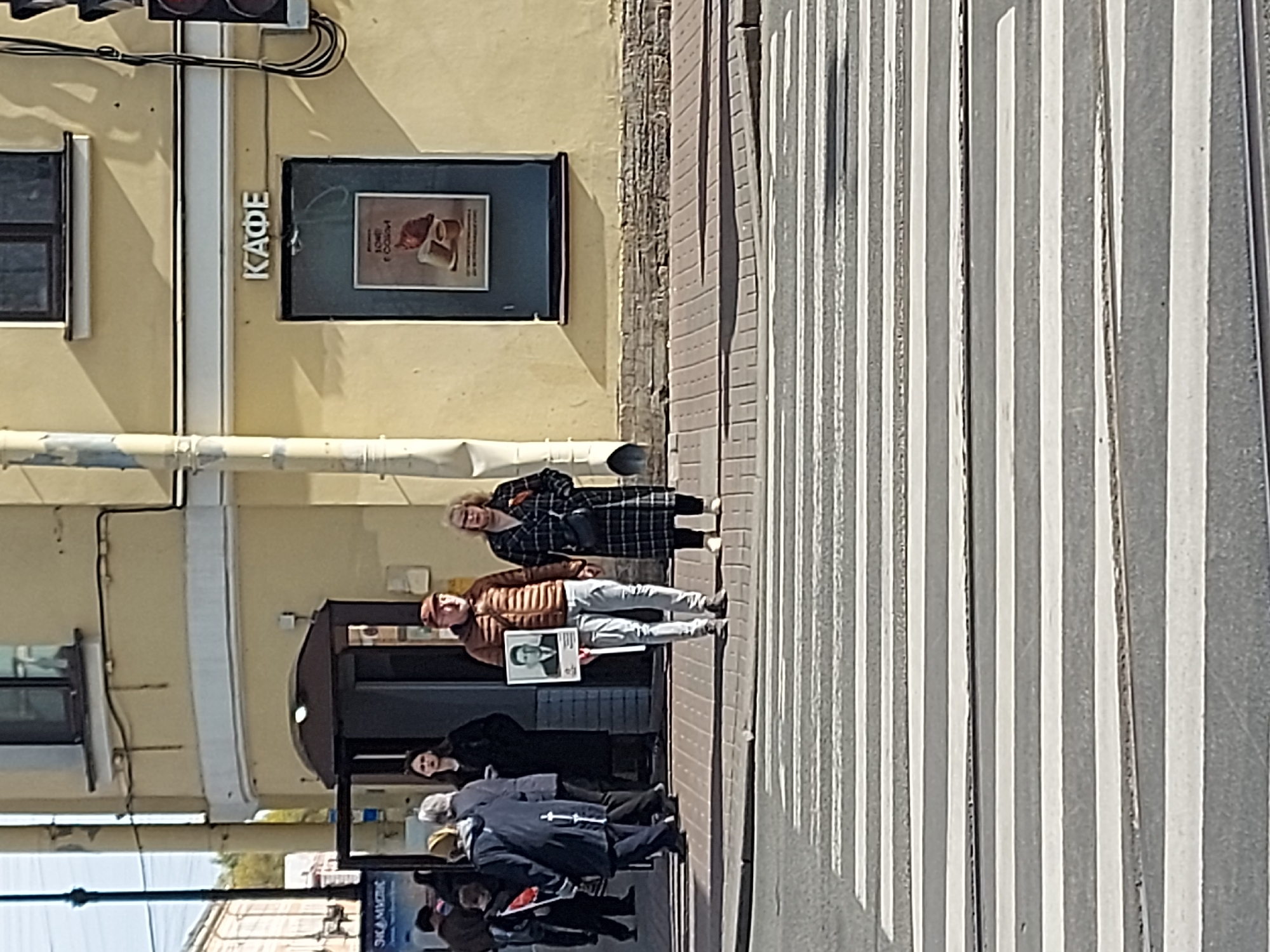 The image shows a group of people standing on a sidewalk in Saint Petersburg, Russia, next to a crosswalk. Several individuals are holding portraits, and one person is wearing a plaid coat. A sign above the entrance reads "КАФЕ" which means "cafe" in English. The building is yellow with a black frame hanging to the right of the cafe sign. There are traffic lights visible on the top left corner of the image.