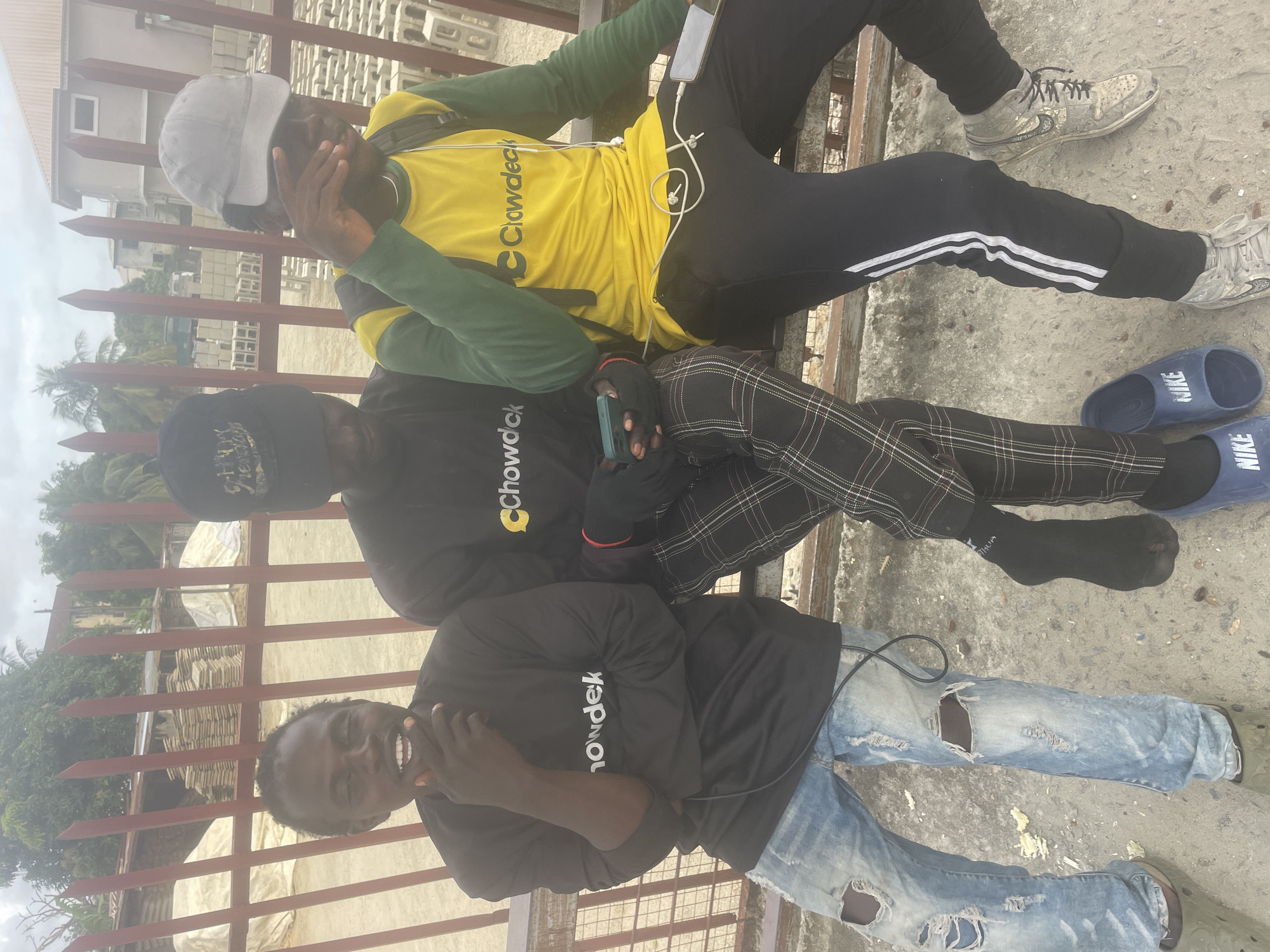 Three young adult males are seated outdoors on a concrete ledge in Ado, Nigeria, positioned in front of a reddish-brown metal fence. The background reveals a construction materials yard with stacks of concrete blocks, wooden planks, and white tarp-covered piles, under an overcast sky. Green foliage and a light-colored multi-story building are visible further in the distance.
From left to right:
1. The first male has dark skin, short dark hair, and is wearing a black long-sleeved shirt with a white "Chowdek" logo. He wears light blue distressed jeans and olive green open-toed slides. His right hand is raised, with index finger and thumb touching his chin, and he smiles towards the viewer. Black wired headphones are draped around his neck.
2. The second male has dark skin, with his face partially obscured by a black baseball cap. He wears a black long-sleeved shirt with a yellow "Chowdeck" logo, black and white plaid trousers, and black socks, paired with blue "NIKE" slides. His legs are crossed, and he holds a teal smartphone in black fingerless gloves, looking down at the device.
3. The third male has dark skin, with his face partially obscured by a grey baseball cap worn backward and his raised left hand. He wears a yellow short-sleeved t-shirt with a black "Chowdeck" logo over a green long-sleeved shirt, black track pants with two white stripes on the side, and worn light-colored high-top sneakers. A black backpack is on his shoulders, and a white smartphone with a connected white charging cable rests on his lap. White wired headphones are visible around his neck and chest.