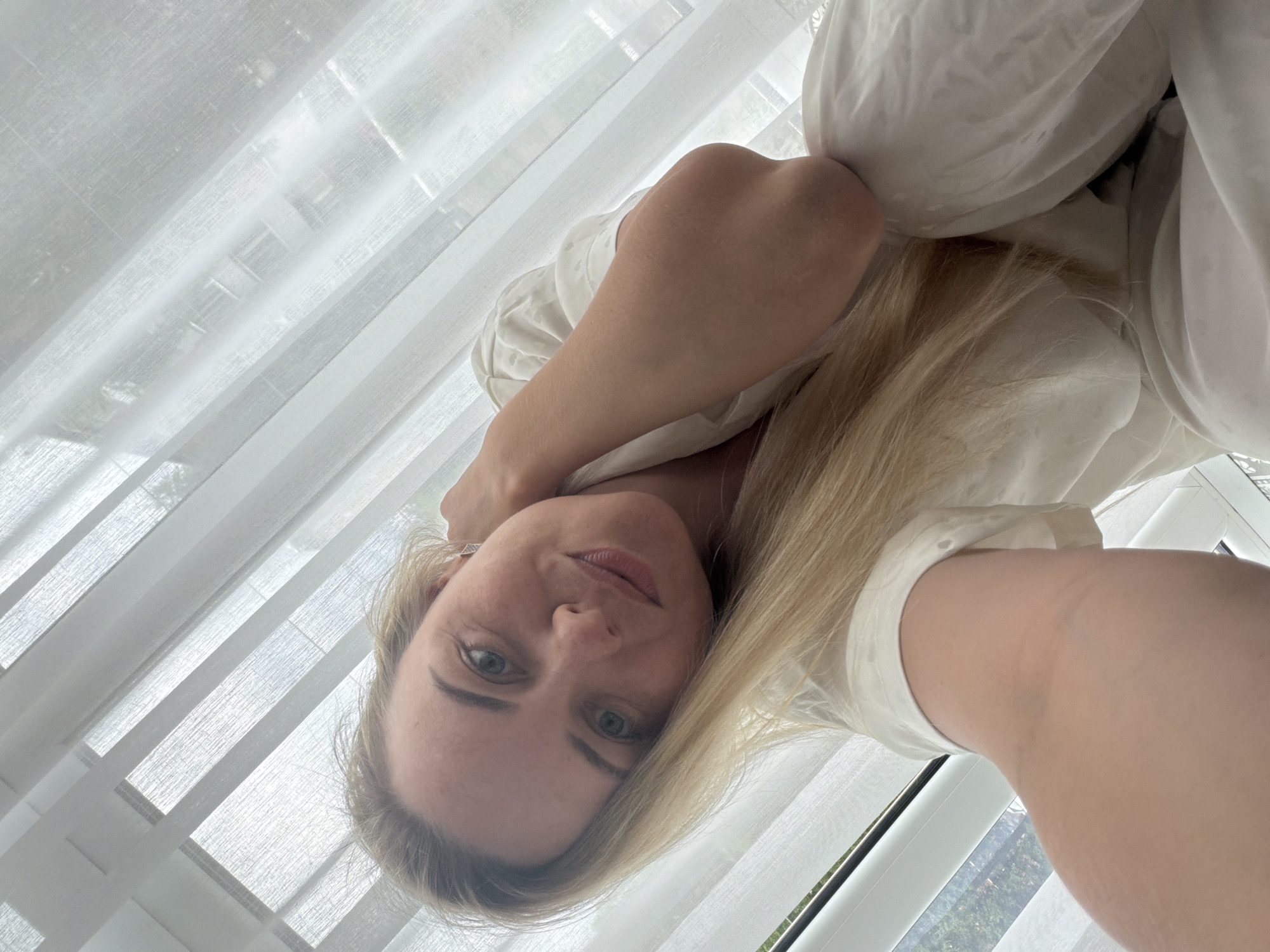 A bright, naturally lit image features a woman taking a close-up selfie indoors, likely in Votkinsk, Russia. The woman, with blonde hair parted on the side and light blue-green eyes, looks directly at the camera with a neutral expression. She wears a white top or dress. A delicate earring is visible in her right ear.
The background is dominated by sheer white curtains covering a large window, allowing soft, diffused light to illuminate the scene. Through the semi-transparent curtains and a gap in the lower right, glimpses of green foliage and outdoor elements can be seen, suggesting a daytime setting with natural light. The woman's arm, visible in the lower right, supports her position, possibly indicating she is lying down or leaning against something soft and white, like a bed or pillows, which are also partially visible. The overall impression is one of calm and natural light. No visible text is present in the image.