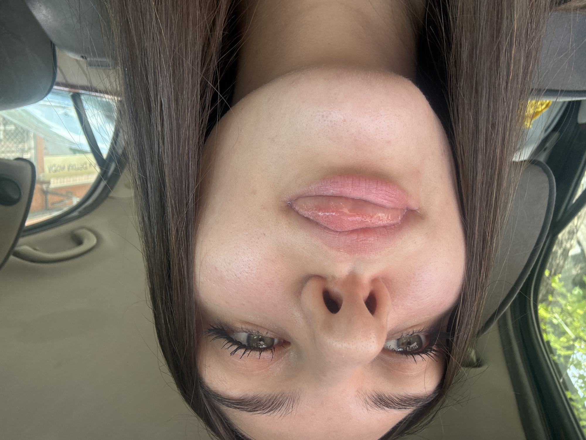 The image, presented upside down, features a close-up of a young woman's face, visible from her nose to the top of her head. She has fair skin, long dark brown hair, and light-colored eyes, possibly hazel or green, with visible mascara on her lashes. Her lips are slightly parted, revealing a portion of her tongue, which appears glossy. Her expression is relaxed and somewhat playful.
The setting is inside a car, with elements like the headliner and window frame visible. Through the car window on the left, an urban or suburban scene is reflected or visible, including part of a building and some green foliage. The time of day is daylight, with ample natural light.
A distinct detail is inverted and mirrored text visible in the reflection through the car window. It appears to read "YDOV AREN," likely from an external sign. Given the provided location context of Sofia, Bulgaria, this could be a distorted reflection of "MODA NOVA" (New Fashion), a common shop name in the region, appearing in a reflected and inverted form due to the image orientation.