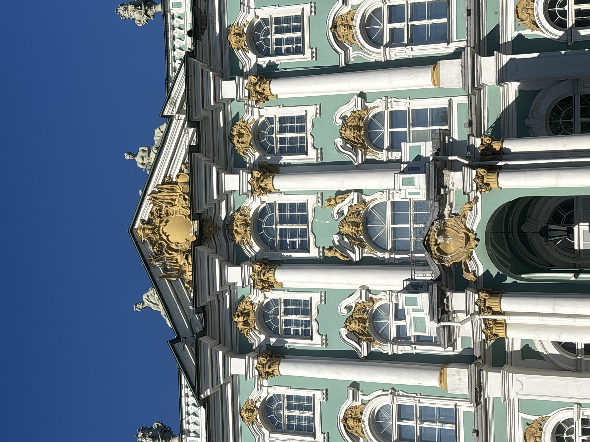 The image displays a close-up, upward view of the ornate mint-green and white facade of the Winter Palace, part of the State Hermitage Museum, located in Saint Petersburg, Russia. The building is illuminated by direct sunlight against a clear, deep blue sky. The facade is characterized by multiple levels of elaborate Baroque architecture featuring numerous white Corinthian columns with gilded capitals and bases, set against a mint-green wall. Arched windows with white frames and multi-pane grilles are uniformly distributed across the visible section, each adorned with intricate gold and white decorative elements, including stylized masks, scrollwork, and pediments. On the roofline, several grey/white statues are visible, including two reclining male figures. A prominent golden escutcheon is centered near the top of the visible facade, below the roofline. Below this, a small balcony or projection is discernible. Towards the bottom center, a large arched entrance or recess is visible, partially obscured. No people or actions are depicted.