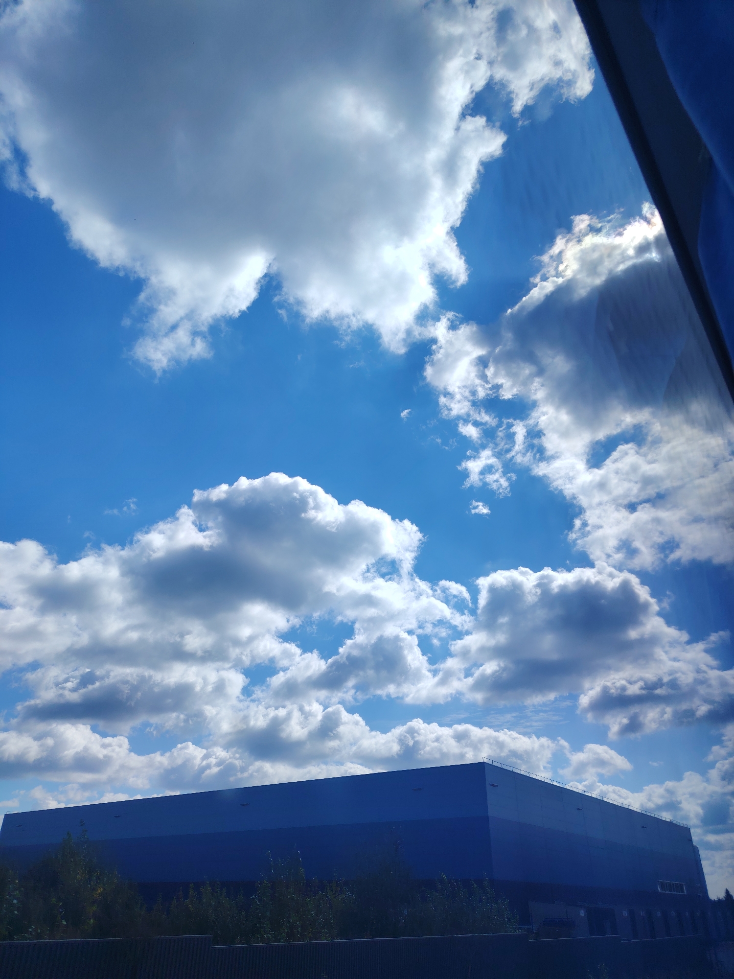 An outdoor, slightly angled, eye-level, full shot captures a large, flat-roofed, dark blue building situated in a landscape with trees and a fence in front of it. Above the building, a vast expanse of bright blue sky is filled with numerous fluffy white cumulus clouds. Sunlight is visible, casting rays and highlighting the edges of some clouds, and a portion of a blue tarp or fabric is visible in the upper right corner of the frame, indicating the photographer may be inside a vehicle looking out a window. The scene suggests a daytime setting with fair weather.