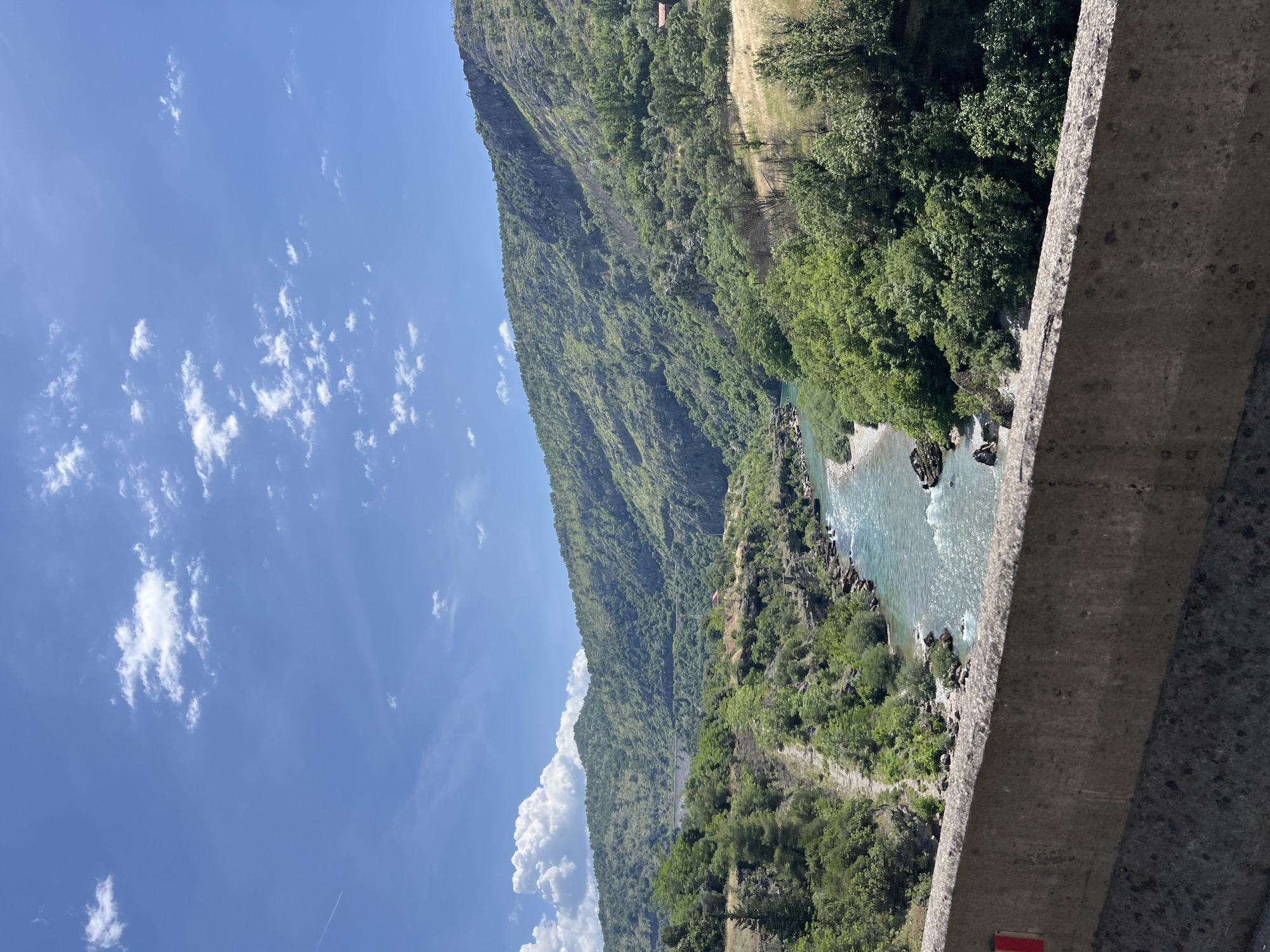 A daytime, landscape view from Donje Mrke, Montenegro, features a river running through a valley at the base of a large, tree-covered mountain. The blue river has areas of rapids and some visible rocks. Lush, green trees and vegetation cover the lower portion of the mountain and line the river's banks. A tall, green mountain rises to the left of the river valley. The top of the mountain features a patch of tan-colored dry grass and a small building. A concrete structure is visible in the foreground. The sky is bright blue with scattered white clouds.