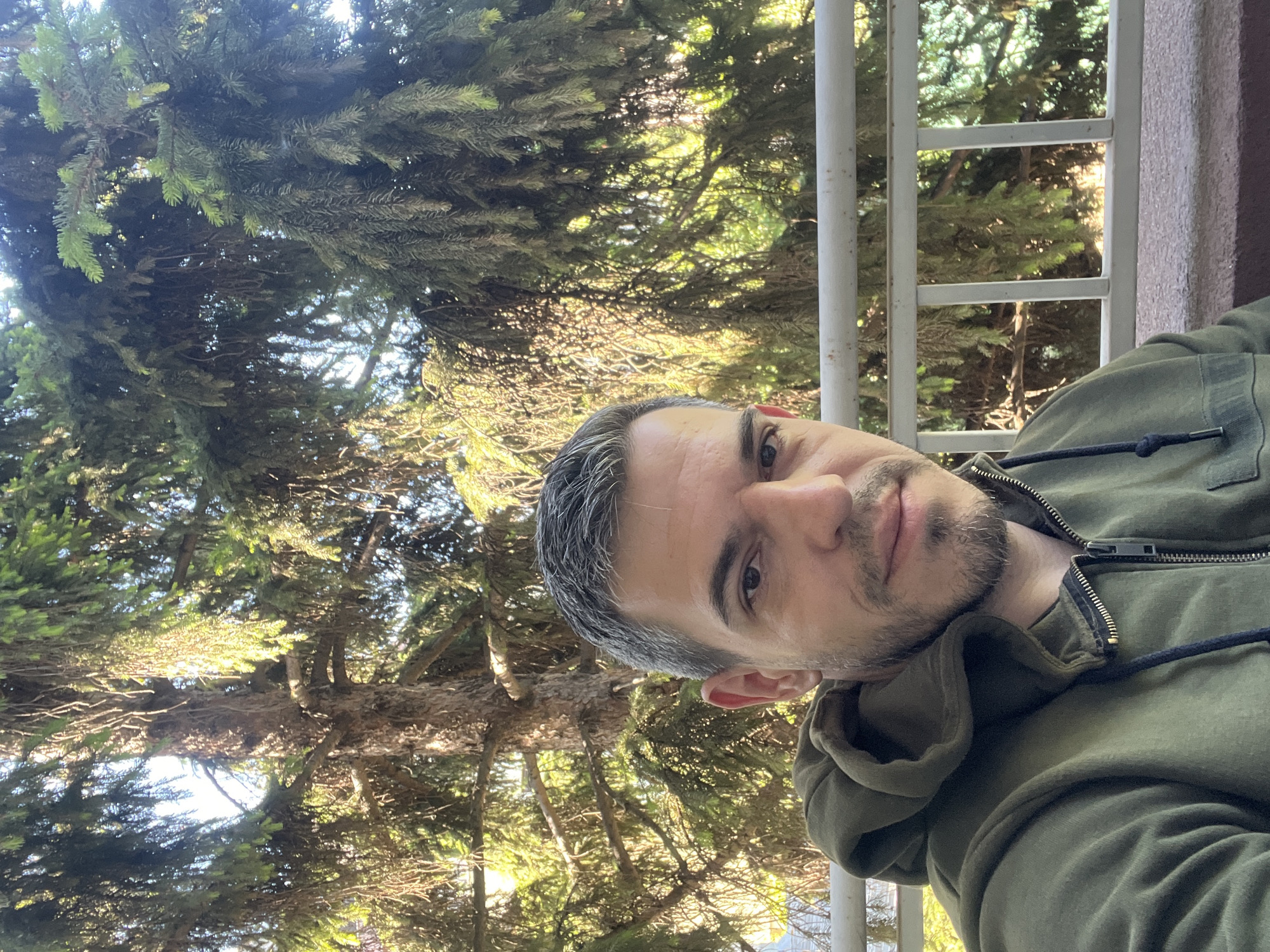 A man with short, graying brown hair and a short beard, wearing a green hooded jacket, is seated outdoors, with a large fir tree in the background and a white railing in front of him. The setting appears to be a balcony or patio in Sofia, Bulgaria, judging by the architecture and vegetation. Sunlight filters through the tree branches, creating a dappled light effect on the scene.
