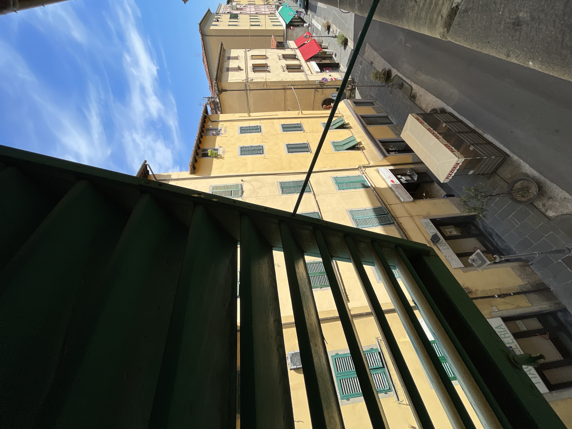 This is a photograph taken from a high vantage point, looking down at a narrow street in Pescia, Italy. The perspective is skewed, with the buildings appearing to lean inward towards the viewer. The scene is dominated by tall, slender yellow buildings with numerous windows, many of which are covered by green shutters. The street below is paved with dark stone and appears narrow, with a few potted plants and streetlights.
On the ground floor of one building, there is a shop with an awning that reads "PIZZERIA PIZZA FOCACCIA" in red lettering. Another sign above it reads "LUCESINI". The sky above is a bright blue with scattered white clouds, suggesting a sunny day. The lighting indicates it is likely daytime, possibly late morning or early afternoon, given the angle of the shadows. The foreground is partially obscured by dark green, slatted structures, possibly part of a balcony or a window covering. The overall impression is one of a charming, old European town with a lively commercial street. There are no people clearly visible in the image.