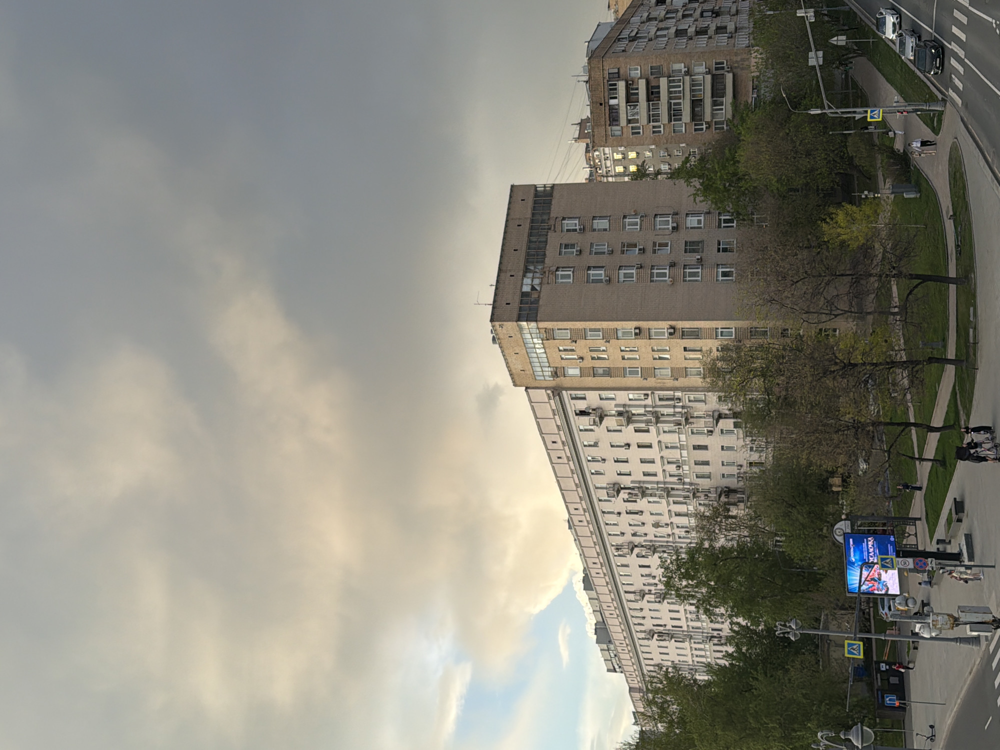 The image shows a city street scene with multiple buildings and trees. The buildings are primarily apartment buildings, with a beige and gray color scheme. The sky is overcast with gray clouds. There are a few people visible on the sidewalk, as well as cars on the street. Streetlights and traffic signals are present, along with a billboard.