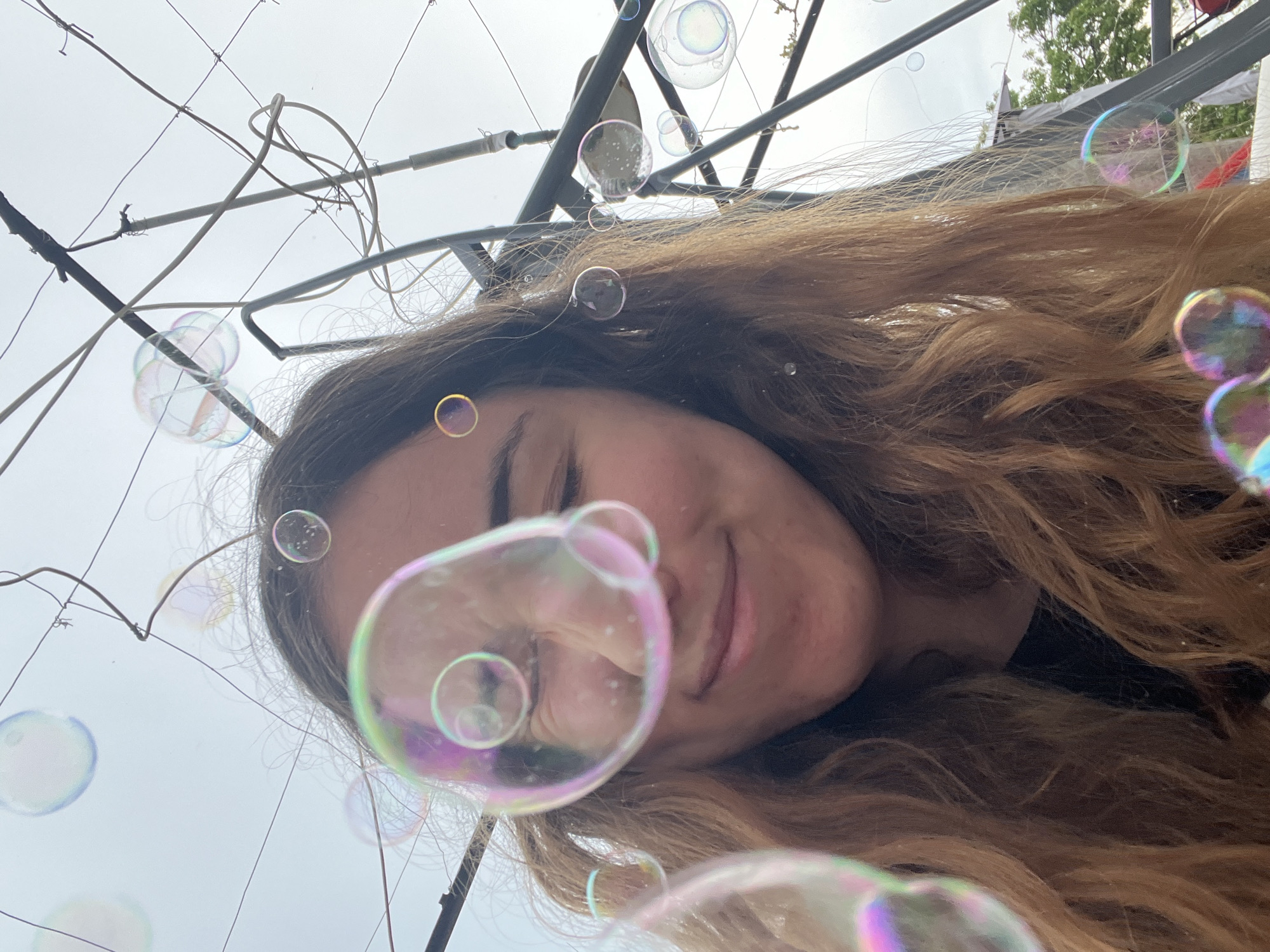 A woman is photographed from a low angle, surrounded by soap bubbles. She has long brown hair and is wearing a black top. The background includes an overcast sky and a metal structure resembling a frame or trellis. Overhead wires and some greenery are visible. There is no direct evidence in the image to confirm that the location is in Burgas, Bulgaria.