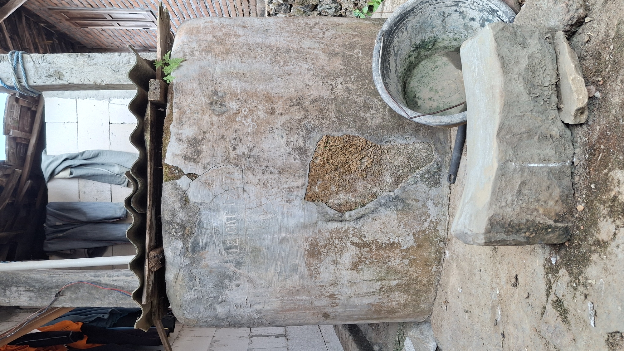 The image shows an outdoor scene in Pucakwangi, Indonesia. In the foreground, a large, weathered concrete water tank is the dominant feature. The tank's surface is rough and shows signs of age, with patches of missing material exposing the underlying aggregate. Etched into the tank's surface are the words "12 OKT 1970". A wavy, corrugated roof made of what appears to be asbestos cement sheeting is supported by wooden beams above the tank. Clothes, including a pair of blue pants and a gray jacket, are hanging from a clothesline strung between a concrete pillar and a wall.
To the right of the water tank, a large, grey plastic bucket is visible, its interior appearing dusty and somewhat stained. A dark, thin rod or handle lies on the ground between the bucket and the tank. In front of the tank, a roughly shaped, light-colored stone object rests on the ground, possibly a makeshift bench or a heavy base. The ground surface is a mixture of dirt, gravel, and scattered debris, with some patches of moss or dampness.
In the background, a brick wall with a wooden-framed window is visible on the right side. A concrete pillar, wrapped with a blue rope near its top, stands to the right of the water tank. A white-painted concrete block wall is partially visible to the left of the tank. The overall impression is one of an utilitarian, outdoor storage area, likely part of a residential or small business property in a rural or semi-rural Indonesian setting. The image is taken from a slightly elevated perspective, looking down towards the scene.