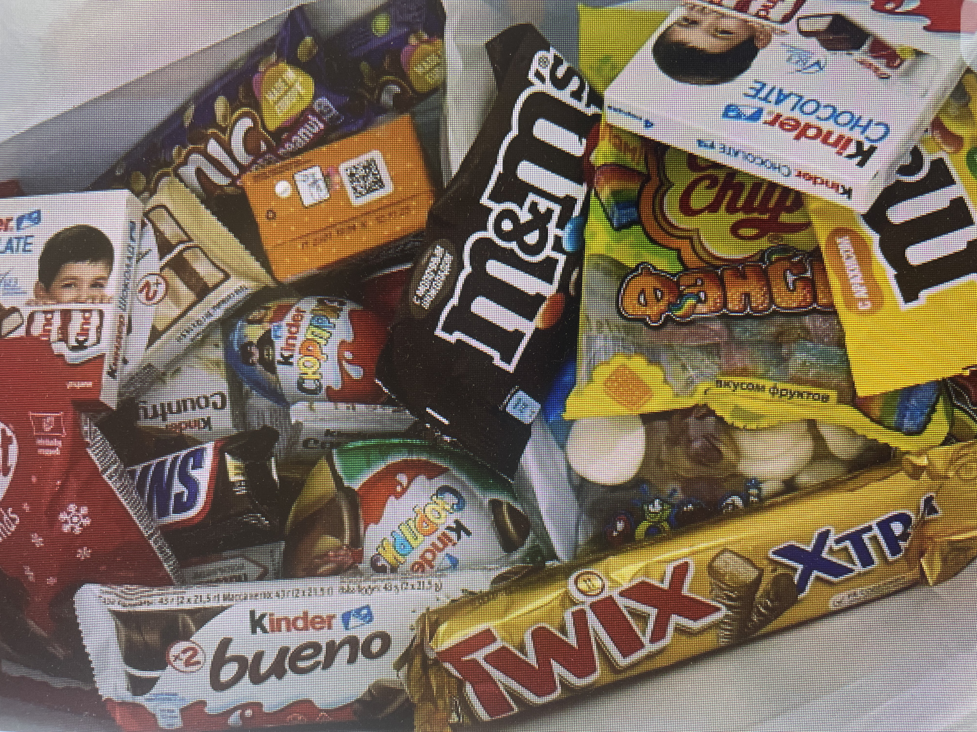 A densely packed collection of various chocolates and candies is presented in a top-down view. The image, exhibiting a noticeable pixelation, appears to be a photograph taken of a digital screen.
The primary subjects are numerous confectionery items from well-known brands. Visible items include multiple Kinder products: Kinder Chocolate bars featuring a child's face, Kinder Bueno (a "2 bueno" pack), Kinder Country, and Kinder Surprise chocolate eggs with the Cyrillic text "СЮРПРИЗ." Other prominent items are a large black-packaged M&M's chocolate, a gold-wrapped Twix bar with "XTRA" visible, and a miniature Snickers bar. A bag of Chupa Chups candy is also present, displaying "Chup Chups" in its distinct logo, along with Cyrillic text "вкусом фруктов" (with fruit flavor) and "ОРЯНЖ" (Orange). An orange-wrapped chocolate bar with a QR code is partially visible, as is a purple chocolate bar, likely from Cadbury, though its brand is not fully legible.
No specific activity or event is taking place; it's a static arrangement of sweets, possibly a gift assortment or a candy haul. The items are arranged closely together, creating a vibrant and appealing display of colors and packaging designs. The mixture of English and Cyrillic text on popular international brands aligns with the provided location context of Poltava, Ukraine.