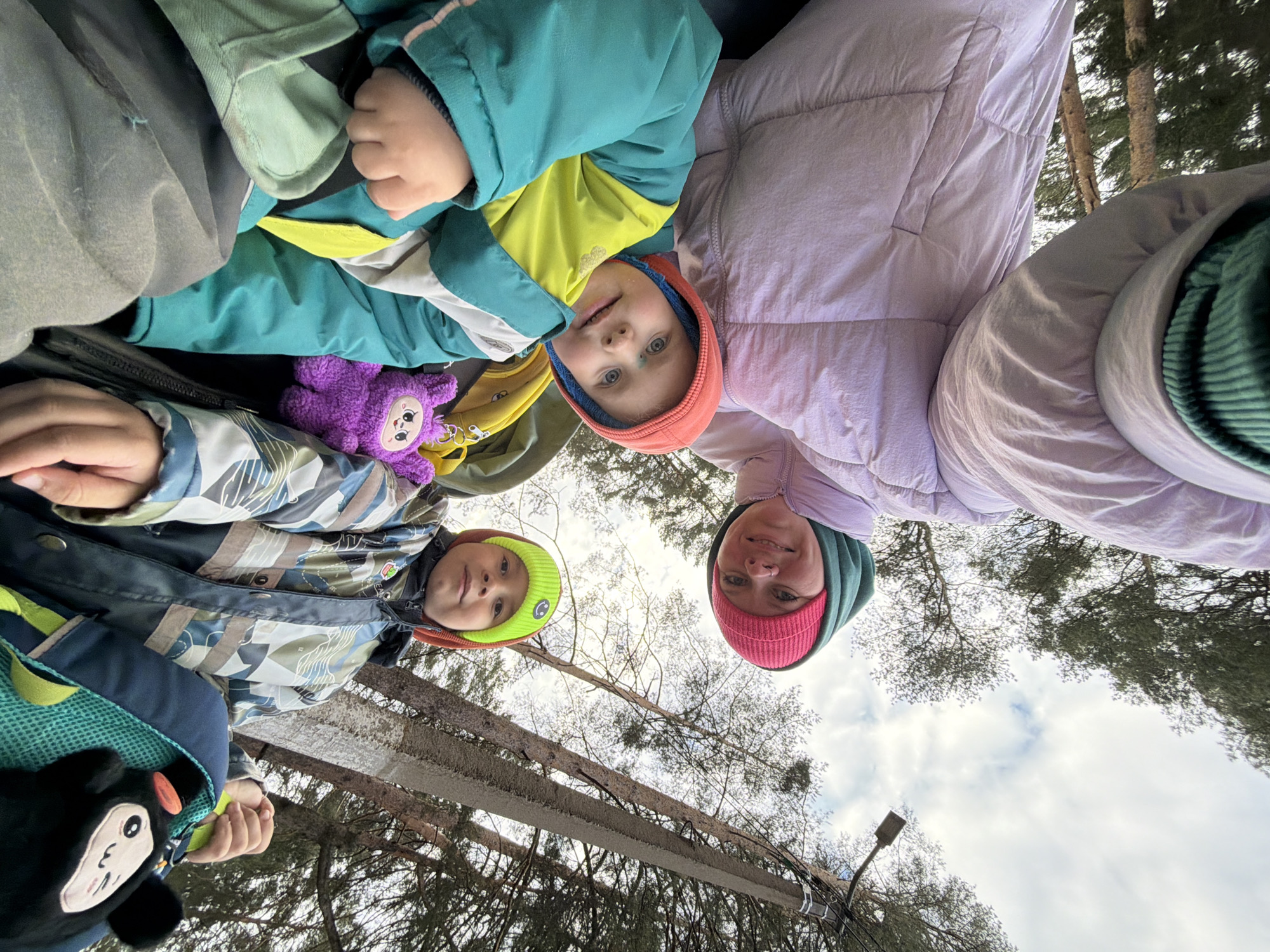 A low-angle shot looking up depicts three individuals and an overhead view of a wooded area. From left to right, the subjects appear as follows: a child in a patterned jacket with a plush toy and backpack visible; another child in a teal and yellow jacket with an orange hood; and an adult wearing a lavender puffer jacket with a pink and green hat. The sky is visible through the tree branches. A street lamp and power lines are situated in the background. The location is the city of Chaikovsky, Russia.