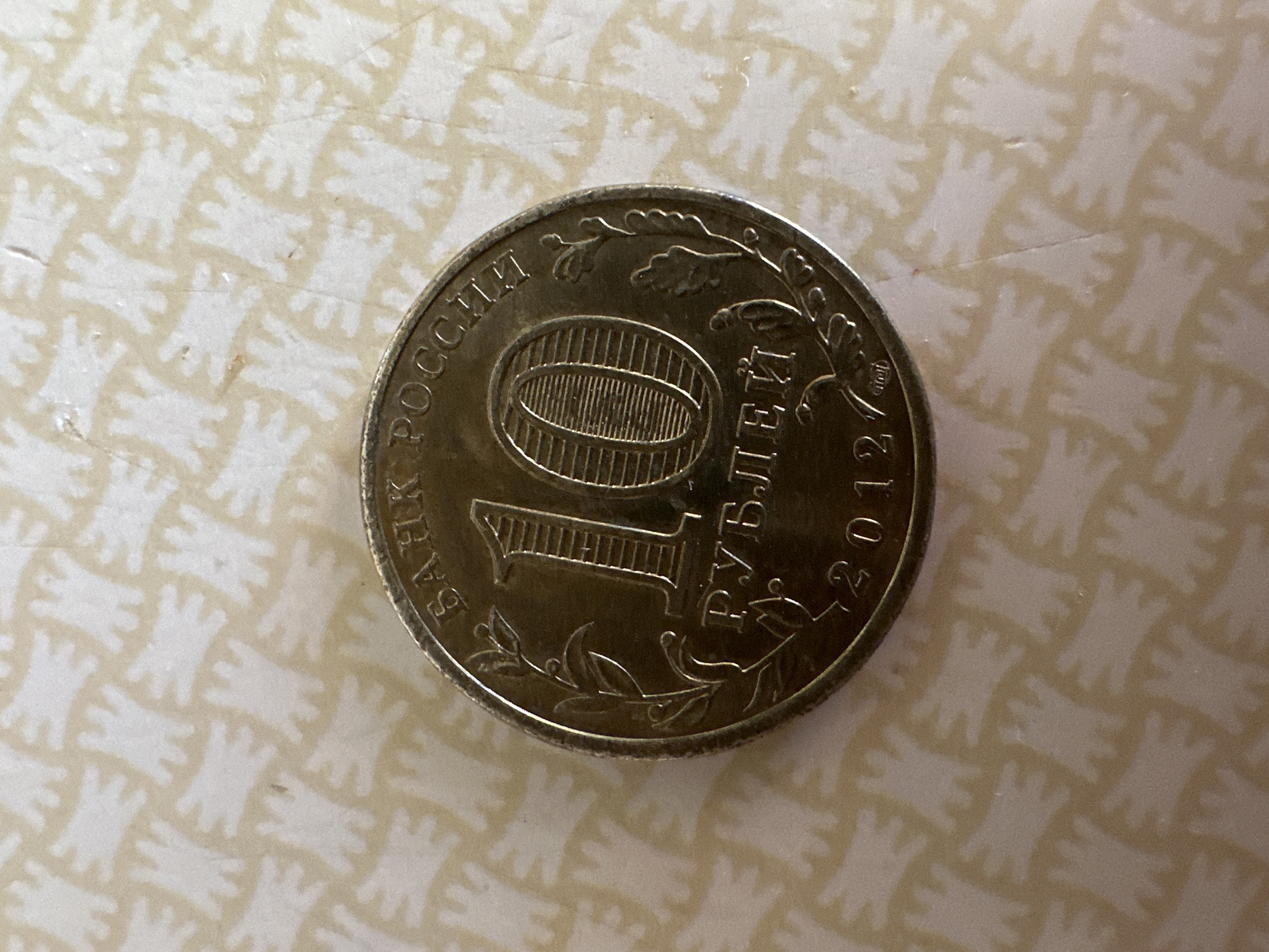 A 2012 Russian 10 ruble coin lies on a light-colored surface with a repeating pattern of stylized, light-colored, animal-like figures. The coin is centered in the frame, oriented with the "10" facing the viewer. The inscription "БАНК РОССИИ" (Bank of Russia) is visible in Cyrillic along the top rim of the coin. Below the "10", the word "РУБЛЕЙ" (Rubles) is also visible in Cyrillic. The year "2012" is inscribed at the bottom of the coin. The coin appears to be made of a brass-colored metal. There are no people or animals visible in the image. The background pattern is uniform and does not provide any specific location information. Therefore, the image does not contain any details that could indicate the coin's location in the city of Biysk, Russia.