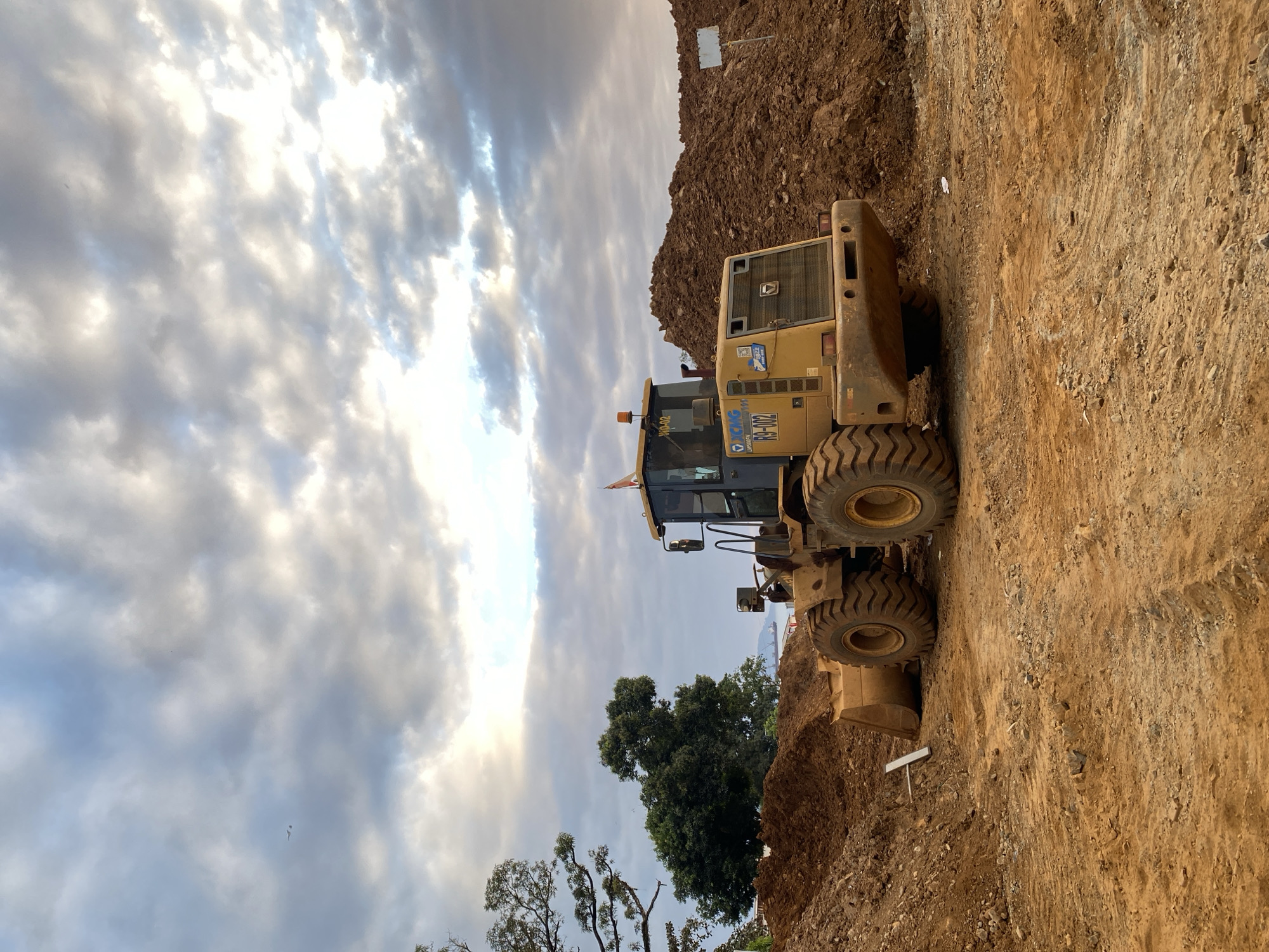 In the city of Unknown, Indonesia, a yellow front-end loader sits idle on a construction site. Piles of dirt surround the machine, and a cloudy sky looms overhead. The loader's bucket is lowered to the ground, and its tires are large and rugged, suggesting it's built for tough terrain. A small flag flies from the cab of the loader, adding a touch of color to the otherwise muted scene. In the background, trees and a body of water are visible, hinting at the natural beauty of the surroundings. The overall impression is one of quiet industry, with the loader poised to resume its work at any moment.