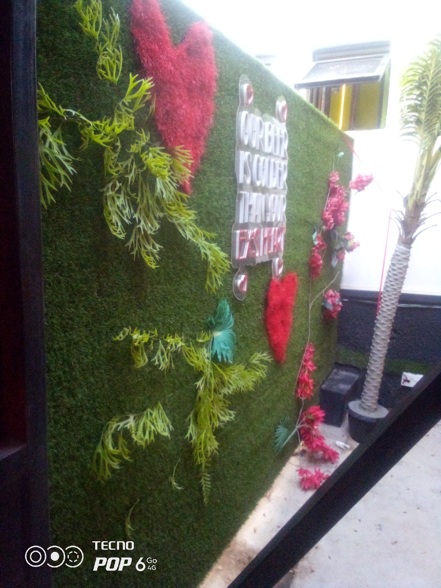 The image depicts an outdoor wall covered in artificial grass, adorned with red heart-shaped decorations and artificial green foliage. A white sign with red trim, containing text in silver and red, is mounted on the wall. The text on the sign reads "OUR BEER IS COLDER THAN YOUR EX'S HEART". To the right of the wall, a portion of a palm tree and a white building with windows are visible. The ground in front of the wall appears to be paved with concrete. A black metal railing runs diagonally across the lower right portion of the image. The bottom left corner of the image shows a logo and text that reads "TECNO POP 6 GO 4G". The scene is likely an exterior decoration for a bar or restaurant in Howayi, Nigeria, designed to be visually appealing and promote a specific product.
