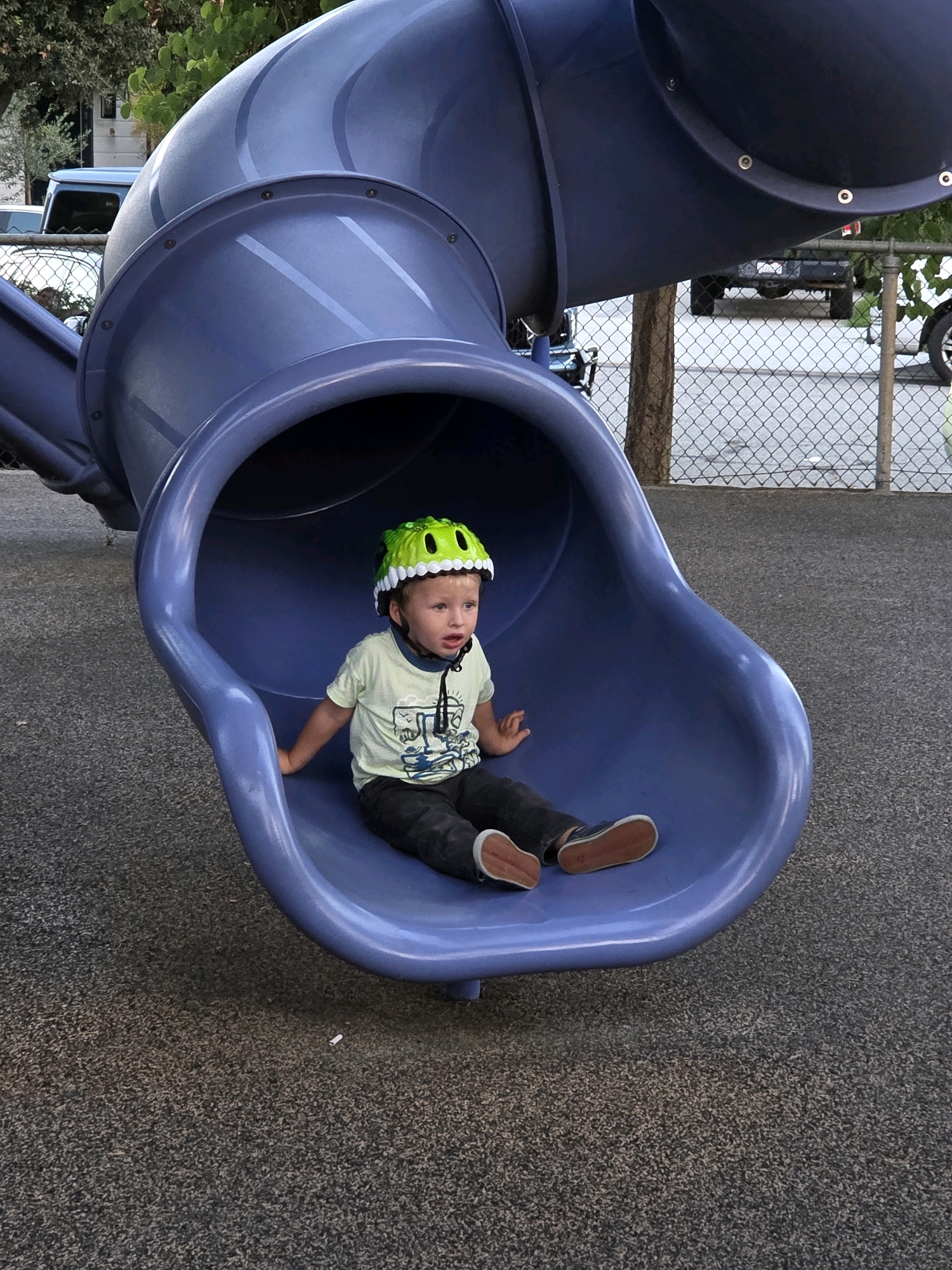 A young boy wearing a green dinosaur-themed helmet sits on a blue plastic slide at a playground. He is wearing a light green t-shirt with a graphic print and dark pants. The boy is looking to his left with his mouth slightly open. The slide is a large, blue, tubular structure. The ground beneath the slide is a dark, pebbled surface. In the background, a chain-link fence is visible, beyond which are trees, a fence, and parked vehicles. The scene appears to be outdoors in a public park.