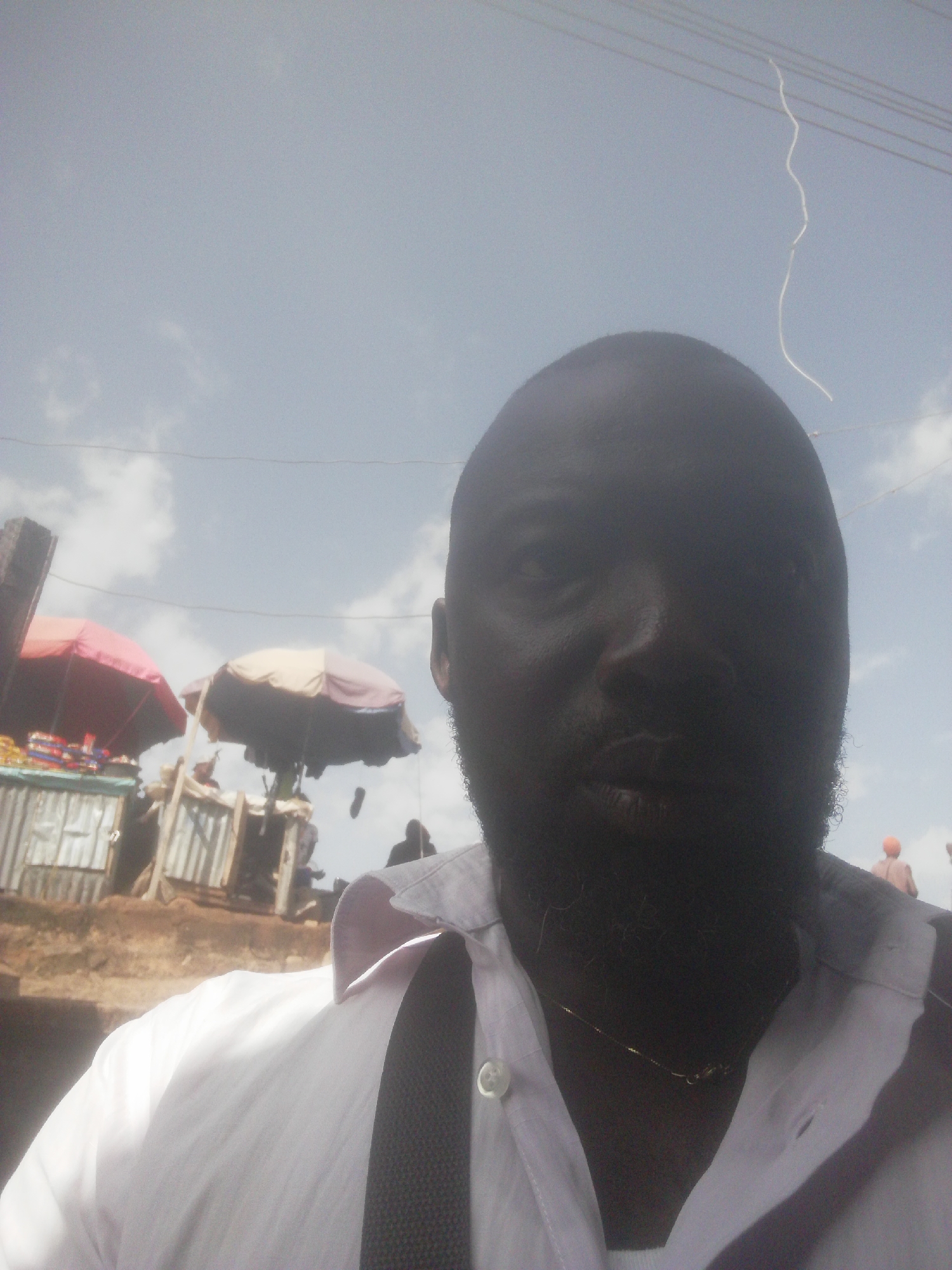 The image is a close-up, waist-up selfie of a man with dark skin, a beard, and a bald head, wearing a light-colored collared shirt and a dark tie. He is positioned in the foreground, with his face largely obscured by shadow. Behind him, the background suggests an outdoor market scene. Two market stalls with umbrellas are visible, one with a red umbrella and the other with a beige and purplish umbrella. In the distance, other figures are faintly visible, along with what appear to be rudimentary structures. The sky above is partly cloudy. The overall impression is that the photograph was taken in a bustling outdoor environment, likely a market in Gwong, Nigeria, on a bright day.