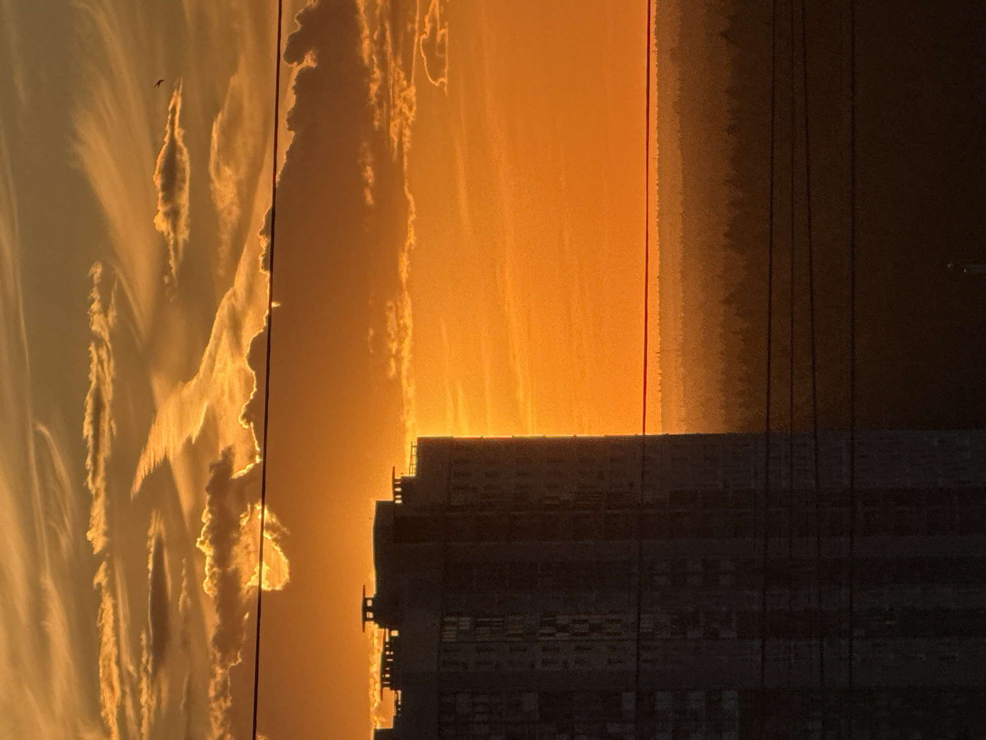 The image shows a silhouette of a tall building against a vibrant sunset sky in Khimki, Russia. The sky is filled with dramatic orange and yellow clouds, creating a striking contrast with the dark, imposing structure. The building appears to be a residential high-rise, with many windows visible, though they are indistinct due to the backlighting. Several power lines run horizontally across the frame, further obscuring the view of the landscape. In the distance, a treeline is visible, forming a dark, layered silhouette against the bright sky. The overall impression is one of an urban environment bathed in the warm, intense light of a setting sun.