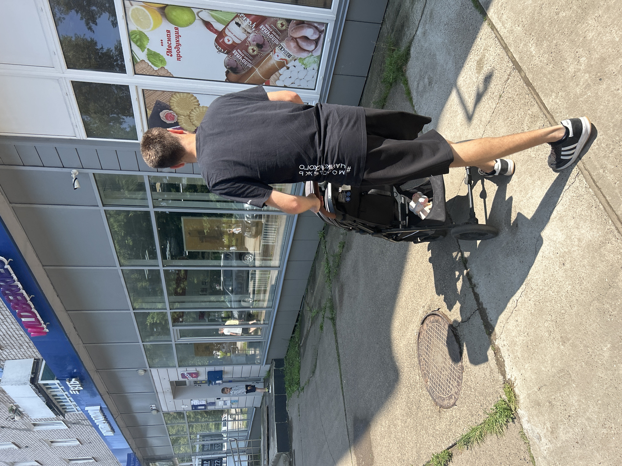 This media file shows a person in a wheelchair on a sunny day, next to a storefront. The person is wearing a black t-shirt with white text that reads "#Молодежь Чайковского", which translates to "#Youth of Chaikovsky". They are also wearing black shorts and black and white athletic shoes. The storefront has large windows displaying images of food items, including sausages, lemons, and chili peppers. Some text on the storefront is in Russian and mentions "мясная продукция" (meat products). The scene appears to be on a sidewalk in front of a building, with grass growing through cracks in the concrete. A manhole cover is also visible on the ground. The sunlight creates distinct shadows on the pavement, indicating it is likely daytime. The overall mood is casual and everyday.