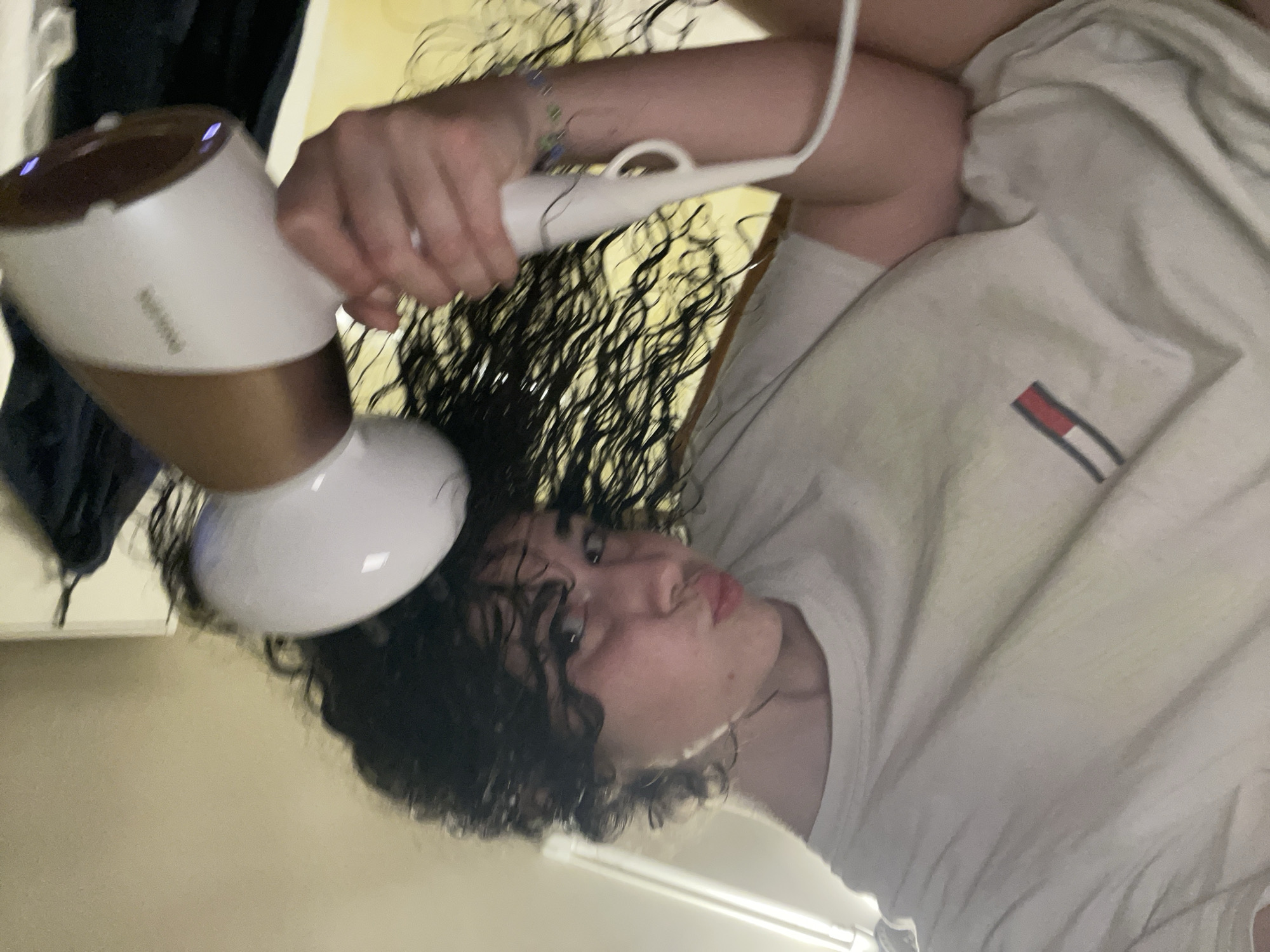 A young individual with dark, wet, curly hair is depicted from a low-angle perspective, drying their hair indoors. The person's head is tilted, and they are looking towards the camera with a slight pout. They are wearing a light-colored short-sleeved t-shirt featuring a red, white, and blue striped logo on the left chest. On their right wrist, a beaded bracelet with green and blue elements is visible. The individual is holding a white and rose-gold Philips hairdryer with a diffuser attachment in their right hand, pointing it towards their wet hair. Blue indicator lights are illuminated on the base of the hairdryer. The background shows a light-colored ceiling or wall with a horizontal light fixture or trim. To the right, dark fabric or clothing hangs on hooks against a light wall. No specific architectural or geographical markers are present to identify the location as Sofia, Bulgaria.