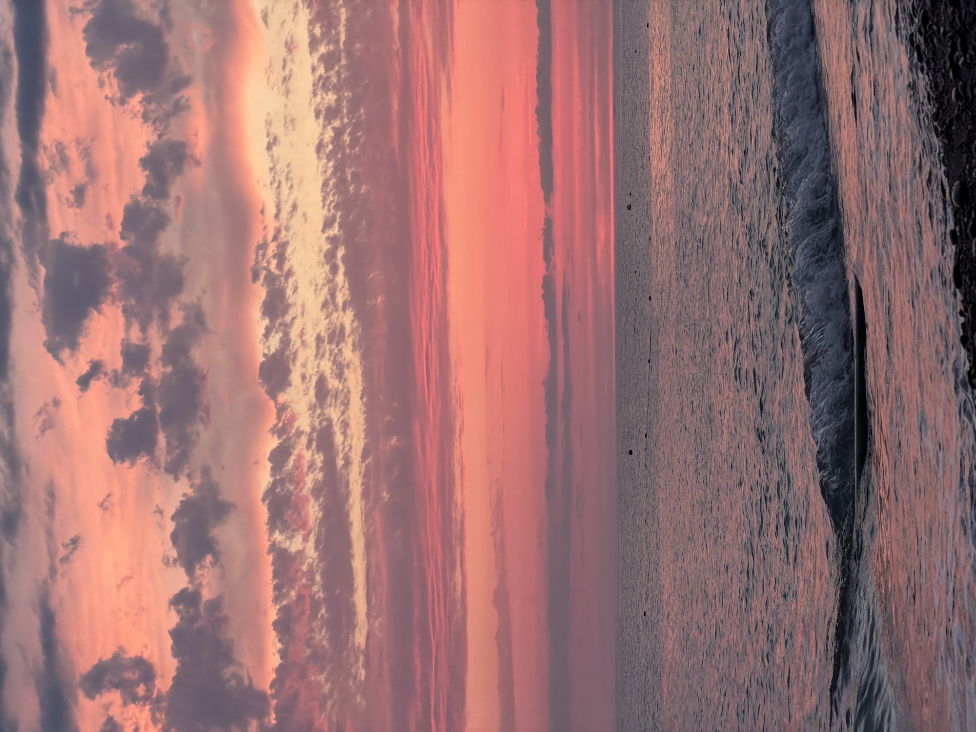An overhead shot shows a body of water with a horizon line, as well as a sky filled with clouds. The body of water is the ocean, and it is calm, with a slight ripple of a wave on the far right of the frame. The ocean is a deep blue, with streaks of pink and orange reflecting from the sky. The sky is filled with clouds, which are tinged with pink and orange from the setting sun. The clouds are of various sizes and shapes, with some being dark and ominous, while others are light and fluffy. The overall impression of the scene is one of peace and tranquility.