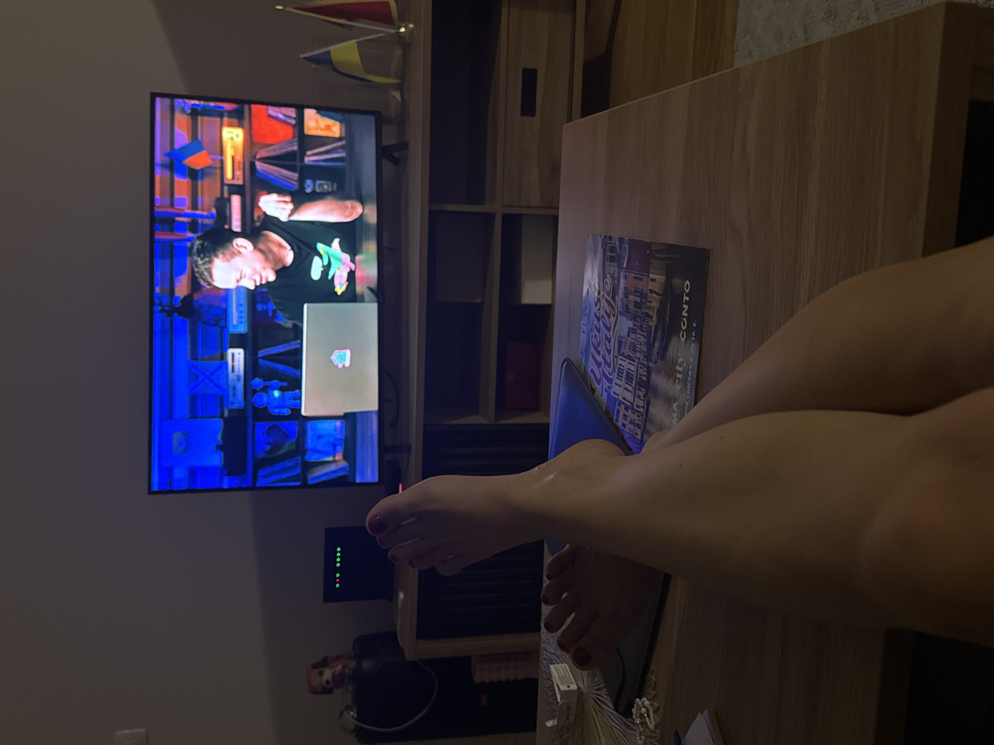 The image shows a person's legs and feet in the foreground, crossed and resting on a coffee table. The coffee table has a magazine on it. In the background, a television is on, displaying a show with a man talking. There are flags on stands next to the television. A shelf unit is behind the television, with books and other items on it. A small dog figurine is visible on a shelf to the left, and a black electronic device with lights is also present. The environment appears to be a living room with dim lighting, suggesting it is nighttime.