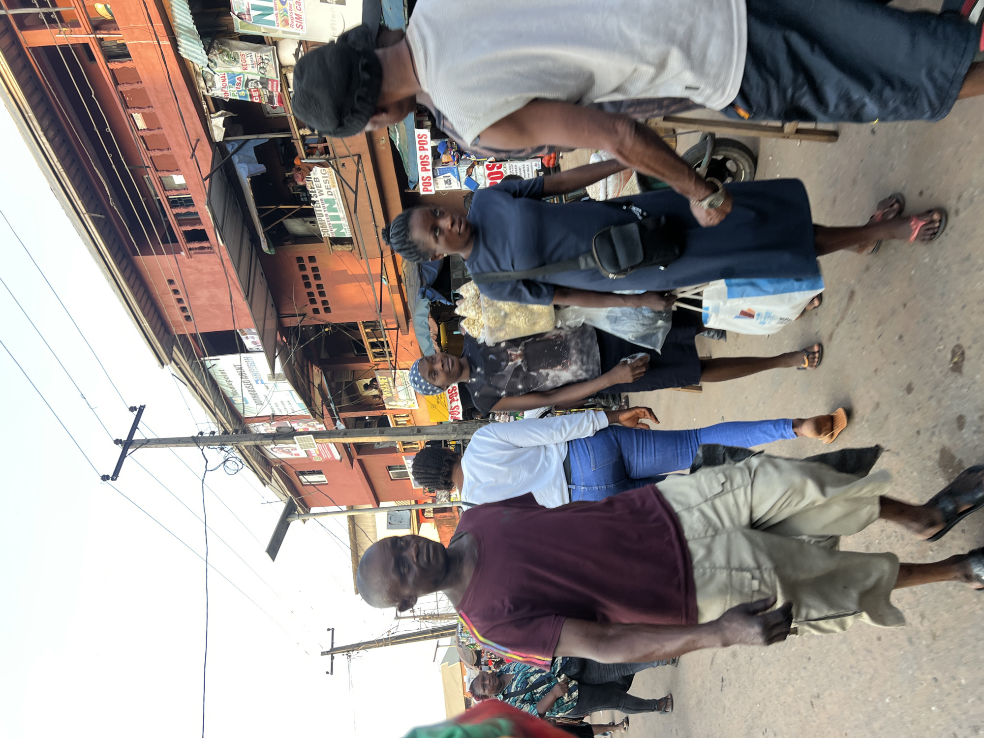 This media file depicts a street scene in Nkpor, Nigeria. Several people are visible, walking along a bustling street lined with buildings and market stalls. The buildings have a mix of modern and older architecture, with some displaying signs for businesses like "Moniepoint" and "Computer Services." The presence of multiple signs advertising "POS" (Point of Sale) services suggests a commercial area.
The people are dressed in casual clothing, and the overall atmosphere appears to be that of a busy marketplace. The time of day seems to be daytime, possibly late morning or afternoon, given the lighting conditions. The sky is bright, indicating clear weather.
One woman in the foreground is wearing a dark blue dress and has a bag slung across her shoulder. Another person is wearing a white shirt and dark shorts. Further down the street, more individuals can be seen, some carrying bags. The ground is paved, and there are power lines and utility poles visible, indicating urban infrastructure.
Notable text in the image includes "POS POS," "Moniepoint," "COMPUTER SER," "REGISTER YOUR WEB," and "NATIONAL IDENTI." These signs provide clues about the commercial activities and services available in the area. The scene conveys a sense of everyday life and commerce in a Nigerian urban environment.
