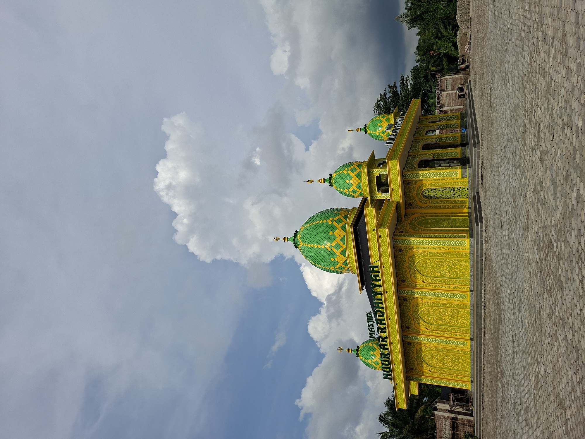 The image shows the Masjid Nour Ar Radhiyyah mosque in Aluekeujruen, Indonesia. The mosque is painted yellow and green, featuring green domes with yellow patterns. The mosque is located on a paved area. The sky is cloudy. The mosque is brightly colored.