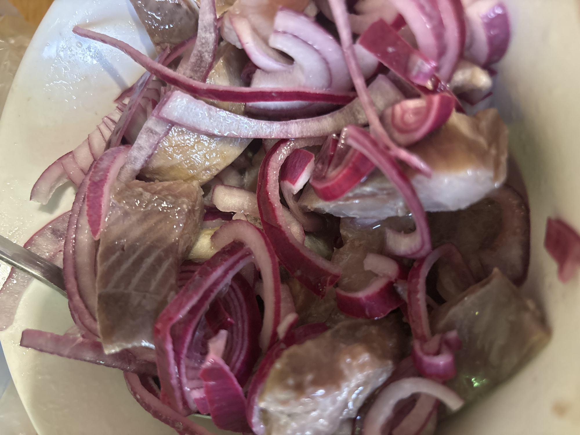 The image shows a close-up view of a bowl containing herring and red onion. The herring appears to be cut into bite-sized pieces and is interspersed with thinly sliced red onion rings. The composition fills the frame, focusing on the textures and colors of the food. The overall scene appears to be indoors, potentially in a kitchen or dining setting. There is no indication of the image being located in Vsevolozhsk, Russia, or containing any people.
