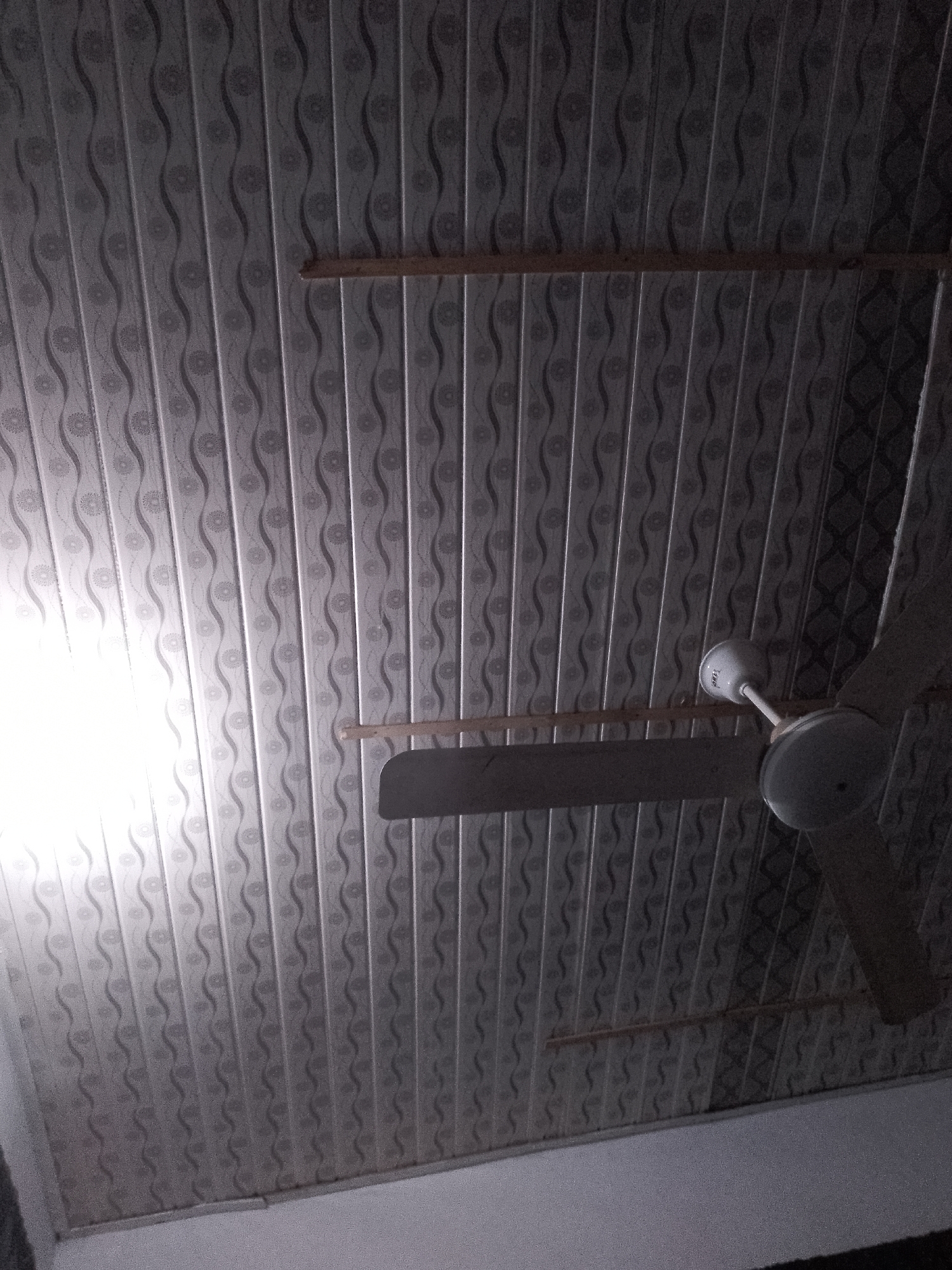 An indoor shot from below captures a ceiling fan suspended from a patterned ceiling. The ceiling features vertical panels with a repeating wavy design interspersed with circular floral motifs. Two wooden beams are visible horizontally across the ceiling, serving as structural supports or decorative elements. The fan itself is white with a central motor housing and three blades, one of which is clearly visible. The lighting in the scene is uneven, with a bright light source on the left side of the image casting a strong glare on the ceiling. The overall impression is of an ordinary interior space, likely a room. The location is not discernible from the image.