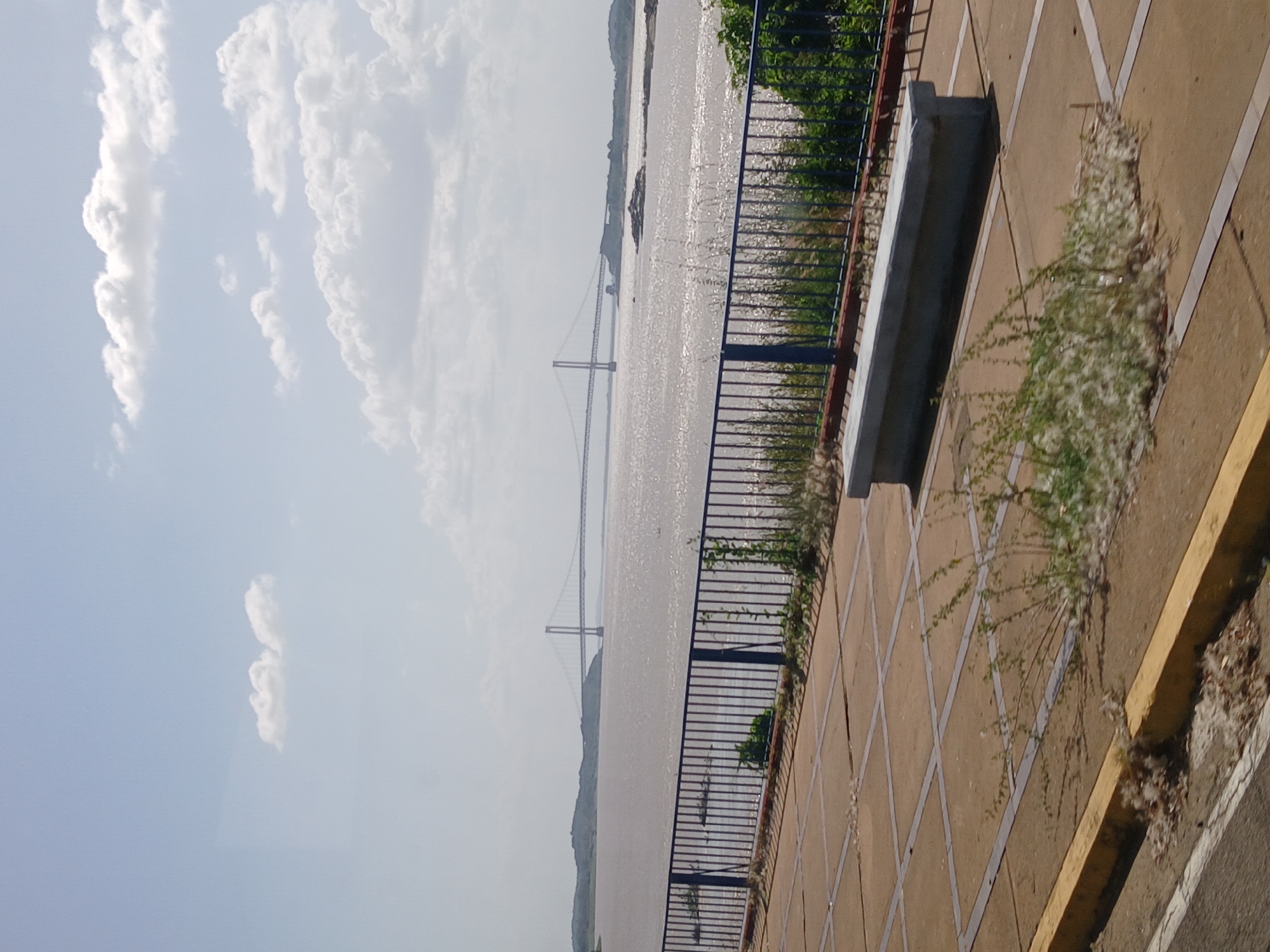 The image shows the Orinoco River in Ciudad Bolivar, Venezuela, with a suspension bridge spanning the waterway. The view is from a raised promenade or walkway along the riverbank, with a low metal railing and a concrete bench separating the walkway from the river. The walkway is paved with square tiles and has some vegetation growing around the edges. The river is wide and appears muddy, reflecting sunlight. In the background, there are landmasses on either side of the river, and the sky is partly cloudy.