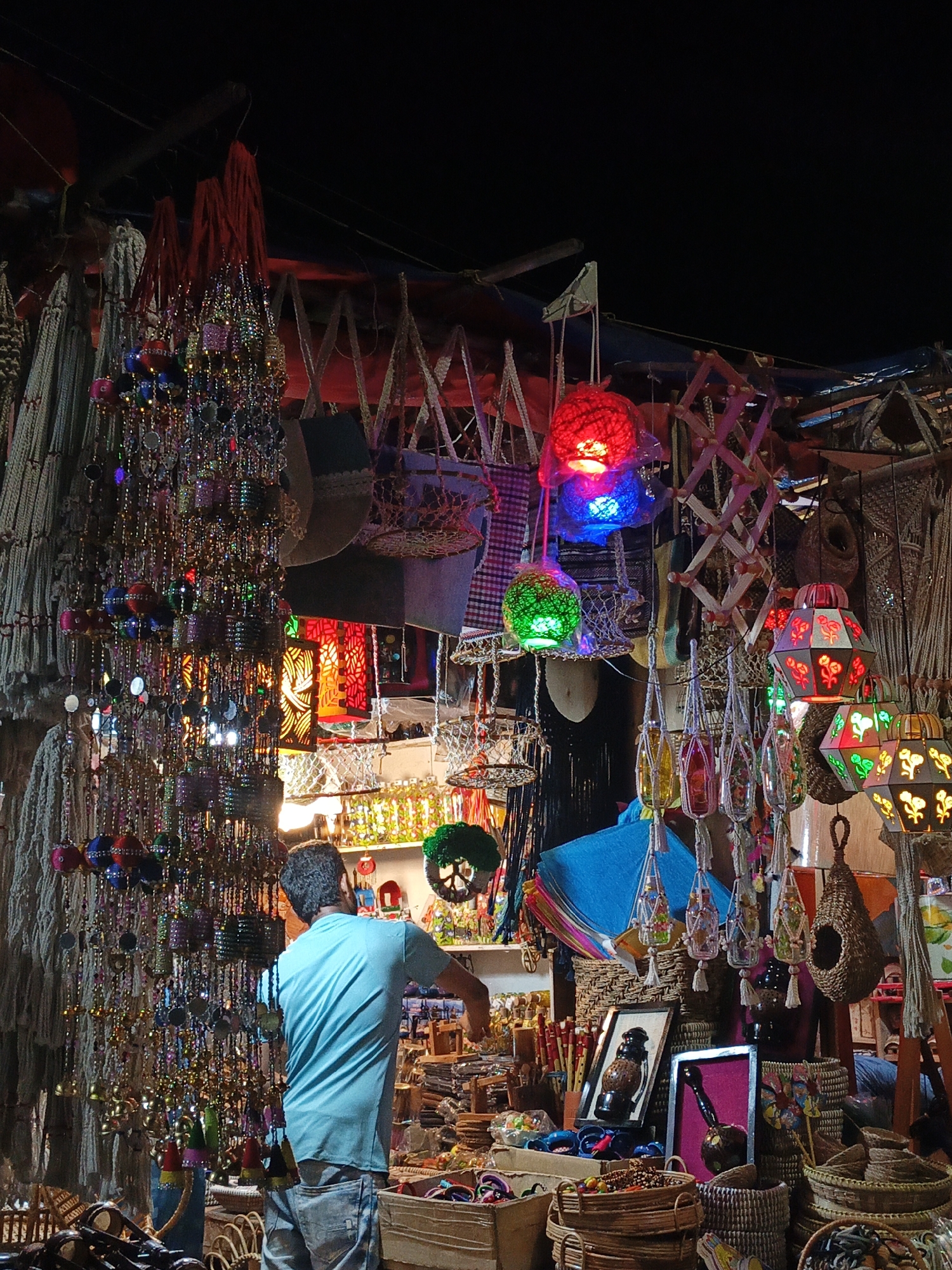 The image presents a vibrant night market stall, likely in Dhaka, Bangladesh, brimming with a diverse collection of colorful handicrafts and decorative items. In the foreground, a man, seen from behind, wearing a light blue t-shirt and jeans, is positioned centrally, appearing to attend to the merchandise or arrange items within the stall.
The stall is densely packed with goods, primarily hanging decorations. To the left, a prominent display features a cascade of glittering strings adorned with beads, small mirrors, and colorful pompoms, creating a sparkling curtain of light and color. Adjacent to these are various woven and macrame items, suggesting intricate traditional craftsmanship.
Above and to the right, the scene is illuminated by an array of hanging lanterns. These include spherical lanterns glowing brightly in red, blue, and green. Nearby, more elaborate, patterned lanterns with intricate cutouts, some featuring floral designs, emit a warm, inviting glow. Other hanging items include small woven baskets, decorative glass bottles, and what appear to be miniature birdhouses.
Below the hanging displays, the stall continues to showcase a wide range of products. Stacks of colorful kites are visible, alongside framed artworks and numerous baskets filled with smaller trinkets, beads, and other handcrafted goods. The artificial lighting, against the dark backdrop of the night, dramatically highlights the textures, colors, and intricate details of the items on display. The overall impression is that of a bustling, lively, and culturally rich market scene. No visible text can be discerned in the image.