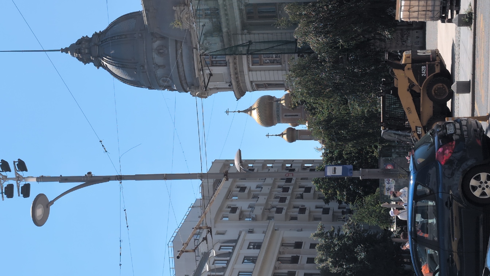 The image depicts a city street scene in Bucharest, Romania, under a clear blue sky. In the foreground, a dark blue hatchback car is parked. Behind it, a yellow skid-steer loader, identified with "CAT 242B" and "SVG," is visible. Further back, several trees line the street.
Dominating the mid-ground is a tall, grey lamppost with multiple lights. Adjacent to the lamppost is a blue bus stop sign. To the right of the lamppost, the golden domes and crosses of a church are prominent, with a larger, ornate grey dome of a building to its right. A multi-story white apartment building stands to the left of the lamppost, featuring numerous balconies. Several people are visible near the bus stop, including two individuals standing together. Power lines crisscross the sky above the scene.