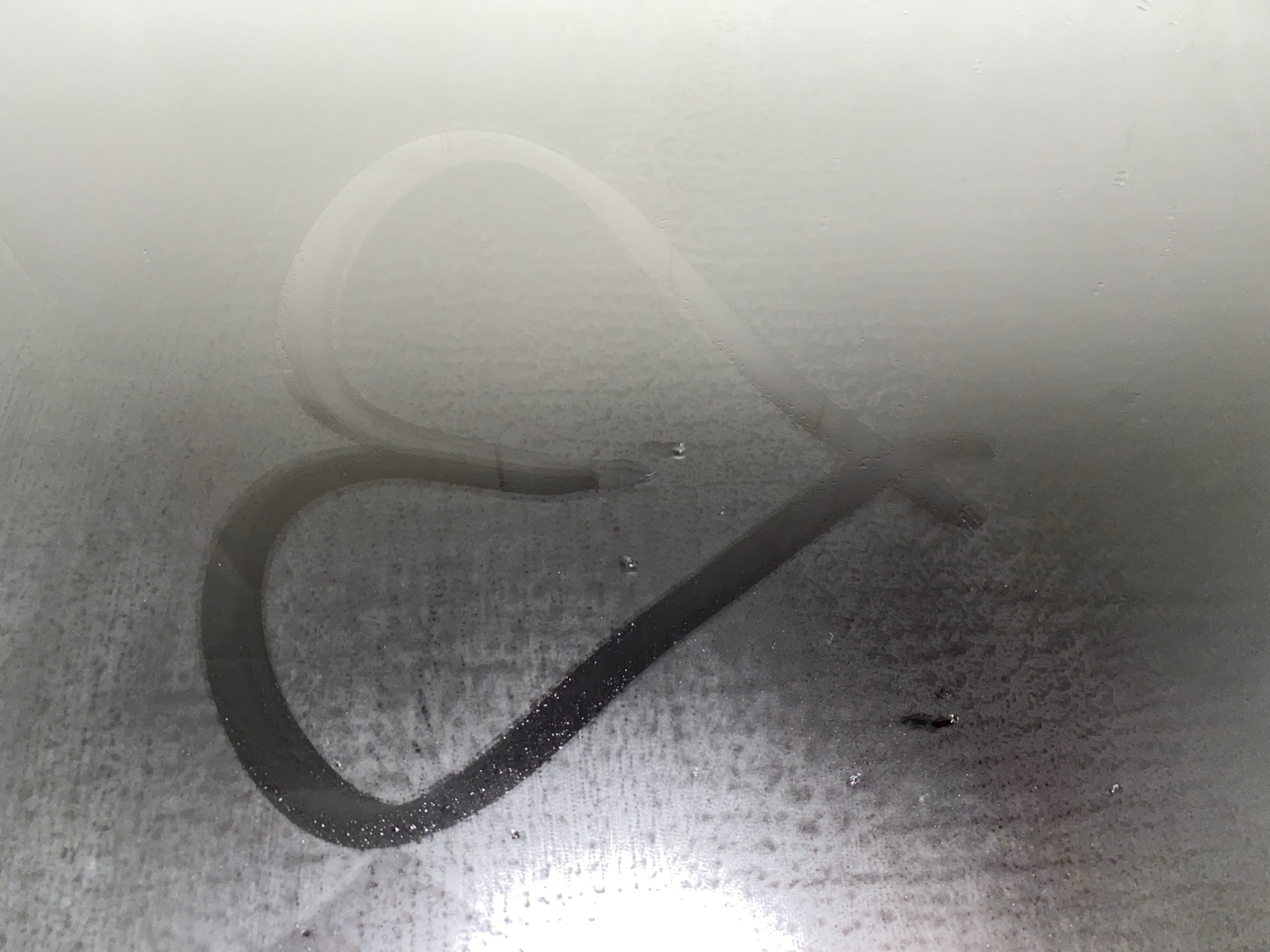 A heart has been drawn in condensation on a foggy surface, likely glass or a mirror. The drawing is in the center of the frame. The surrounding surface is covered in a film of moisture, heavier at the bottom and thinning out towards the top. There are a few visible water droplets in and around the heart. The lighting is diffuse and soft, suggesting an indirect light source. No indication of the image being in the city of Votkinsk, Russia. No people are present.