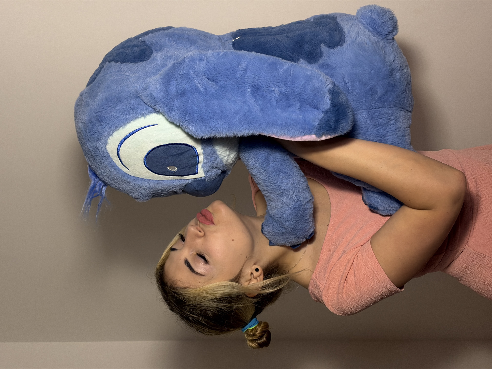 An image, rotated 90 degrees clockwise, captures a young woman affectionately holding a large blue plush toy resembling Disney's Stitch. The woman, positioned towards the right side of the frame, has blonde hair styled in a bun with a blue and green scrunchie. She wears a textured light pink or coral-colored dress. Her eyes are closed, and her lips are slightly pursed, conveying a sense of tenderness and contentment as she holds the furry toy close to her face.
The plush toy, a prominent subject, is a sizable rendition of Stitch, featuring its characteristic blue fur, large white and dark blue eyes, and snout. It appears soft and well-loved. The activity depicts a personal and gentle interaction, suggesting a moment of comfort or affection.
The background consists of a plain, light-colored wall, indicating an indoor setting. The lighting is soft and even, without harsh shadows, implying either diffused natural light or gentle artificial illumination. No text is visible in the image. The overall mood is peaceful and intimate. This scene is located in Budva, Montenegro.