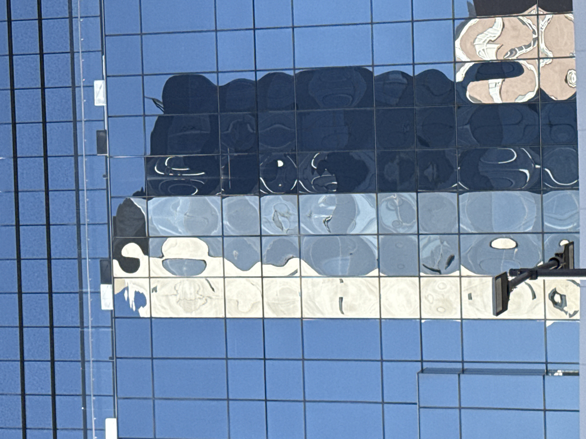 The image captures a portion of a glass building facade in Puebla, Mexico. The building's exterior consists of numerous square panels arranged in a grid pattern. Reflections on the glass surface reveal distorted images of nearby structures and elements.
A building exterior is reflected prominently, visible in the upper portion of the glass panels. Lower panels have reflections that are lighter in color.
To the right side of the image, a mechanical arm extends out from a building, indicating the presence of maintenance or utility equipment.