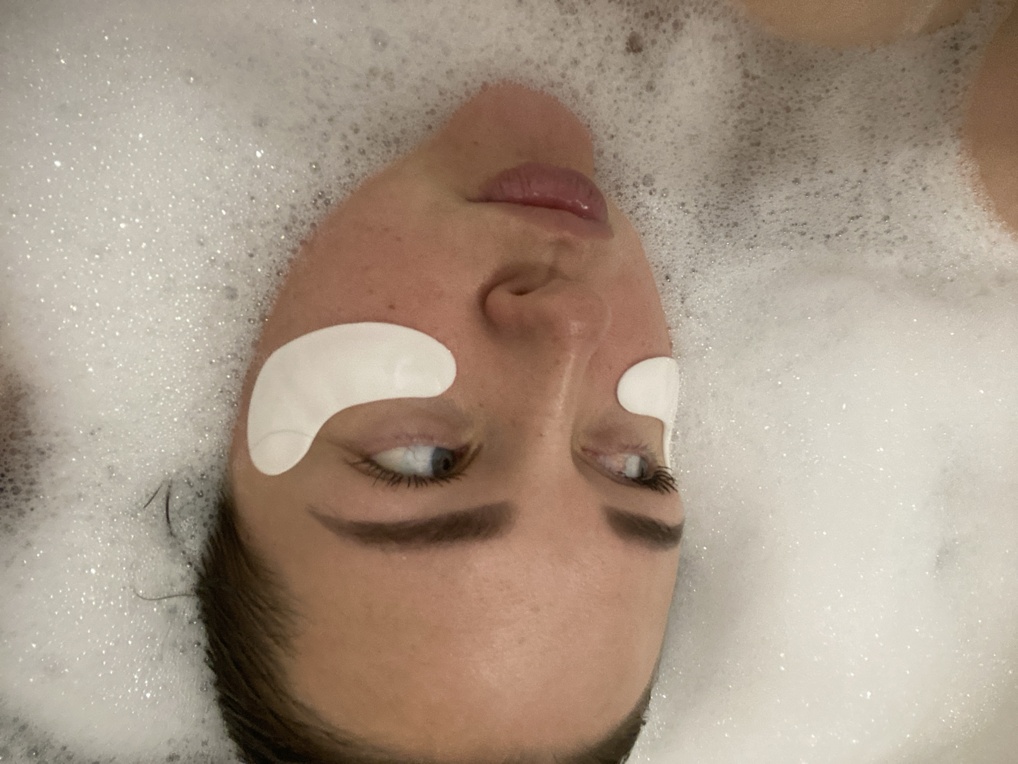 A woman is lying in a bathtub filled with bubbles. She has white eye patches under her eyes. Her face is looking to the side. Her hair is dark and wet. The background is filled with white bubbles. The image does not contain information to confirm that this picture was taken in Sochi, Russia.