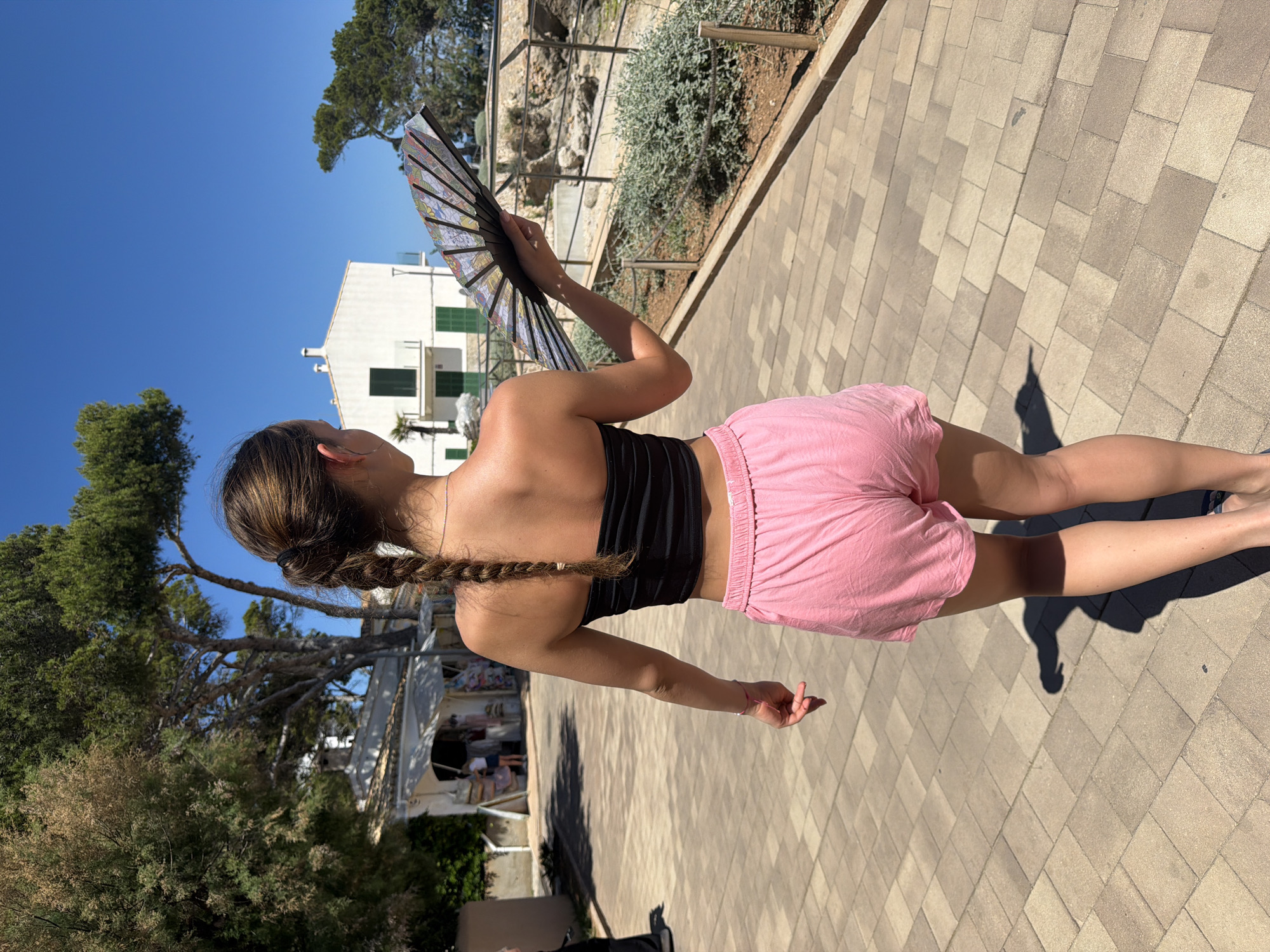 The image shows a person, presumably female, standing on a paved pathway. The person is wearing a black top and pink shorts, with her hair in a braid. She is holding an open fan in her right hand. In the background, there is a white building with green window shutters. A low stone wall with vegetation behind it is also visible. Trees are present on either side of the image. The clear blue sky suggests it is a sunny day. The location is likely within the city of Capdepera, Spain, judging by the architectural style.