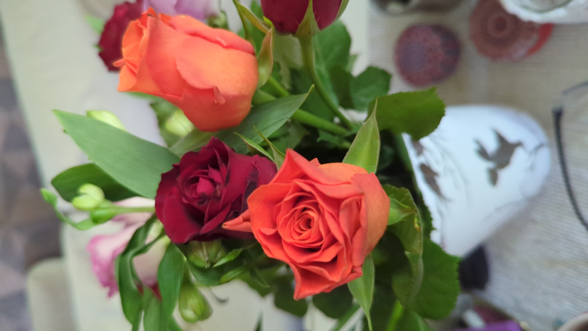 The image shows a close-up of a floral arrangement, primarily featuring several roses. There are two prominent orange roses, one in the foreground and one slightly higher and to the right. A deep red rose is visible behind and to the left of the foreground orange rose. There are also hints of pink and purple flowers or foliage in the background, as well as green leaves and stems. The arrangement is in a white vase with a decorative pattern that appears to depict stylized birds or animals. The vase rests on a textured surface, likely a tablecloth or runner, which is a light neutral color. In the background, on the right side of the image, there are two circular objects, one reddish-purple and textured, the other a darker reddish-brown with concentric circles. The lighting is soft, suggesting an indoor setting. No people are visible in the image. The location in Kløfta, Norway, is not discernible from the visual content of the image.
