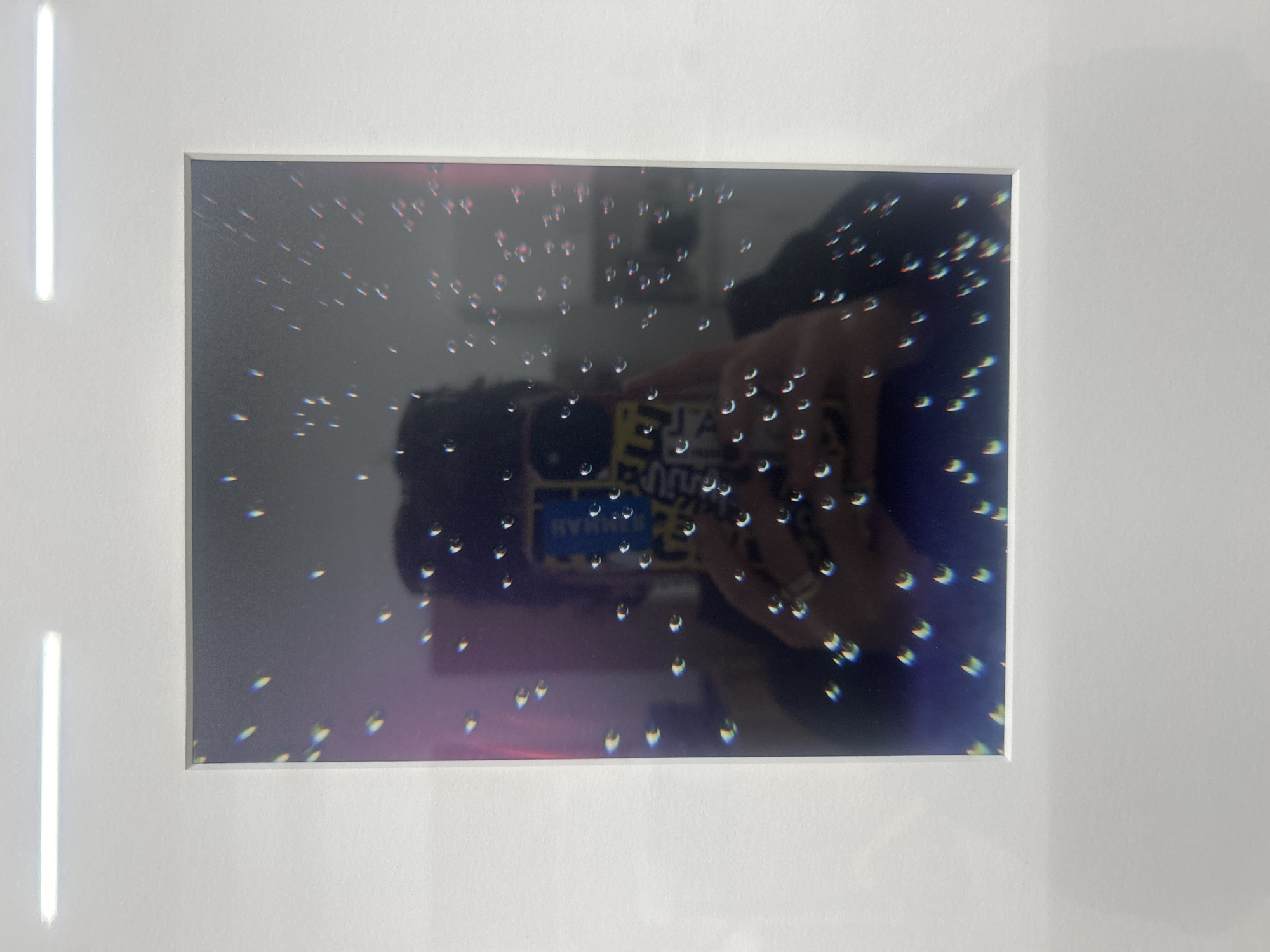 A rectangular framed image shows a photograph of a person holding a phone. The phone reflects the person's face and clothing. The photograph contains small, distorted, colored reflections. The framed image has a white border. The location cannot be determined from the image.