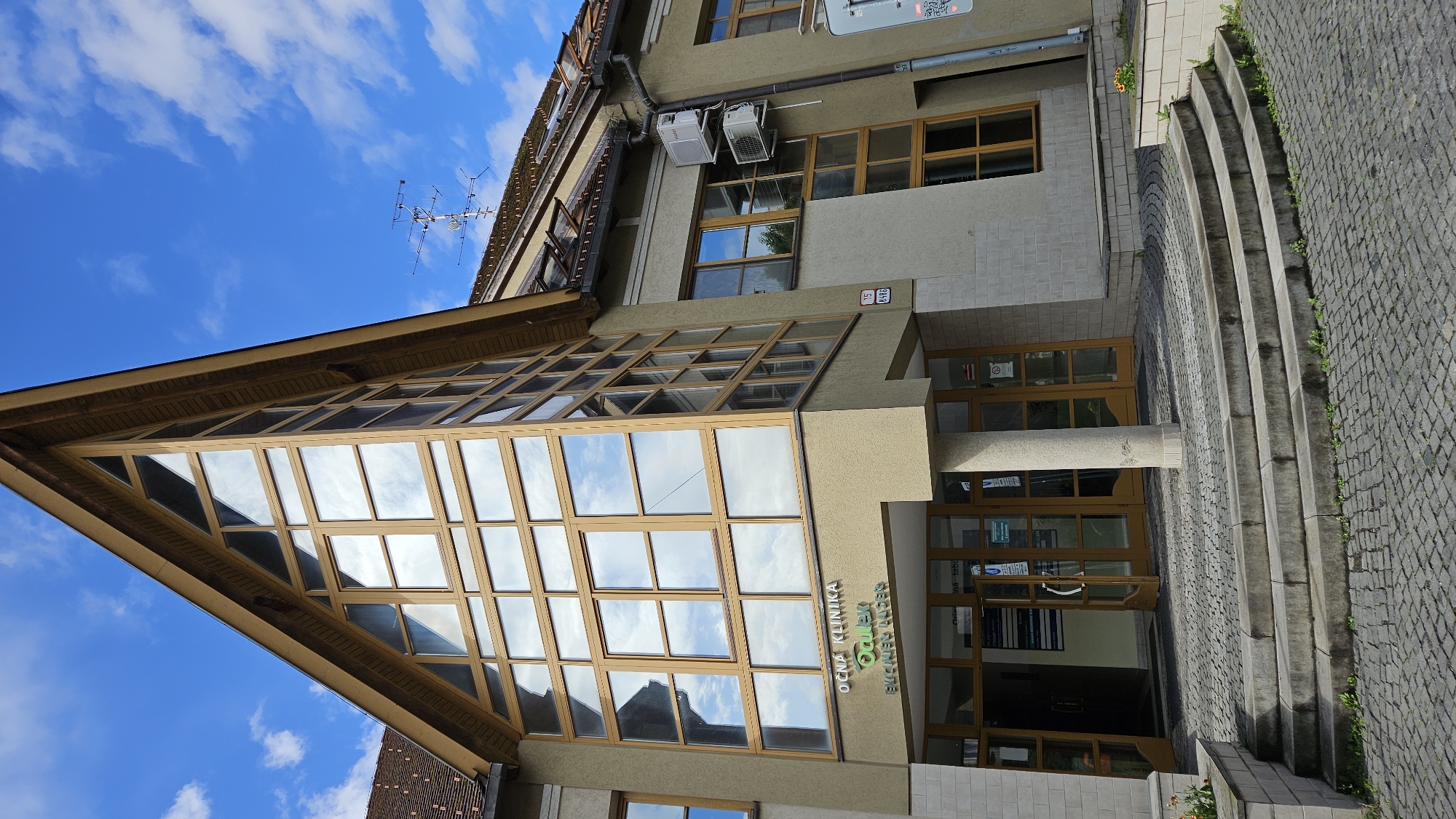 The image depicts the exterior of the Očná klinika Okular in Prešov, Slovakia. The building features a distinctive triangular facade with a large, multi-paned window structure. The clinic's name is visible on the facade above the entrance. The entrance itself is framed by glass doors and a stone column. Paved steps lead up to the entrance area, which is surrounded by cobblestone paving. The sky is blue with scattered clouds, and a rooftop antenna is visible on an adjacent section of the building. Two air conditioning units are mounted on the exterior wall. The scene is brightly lit by daylight.