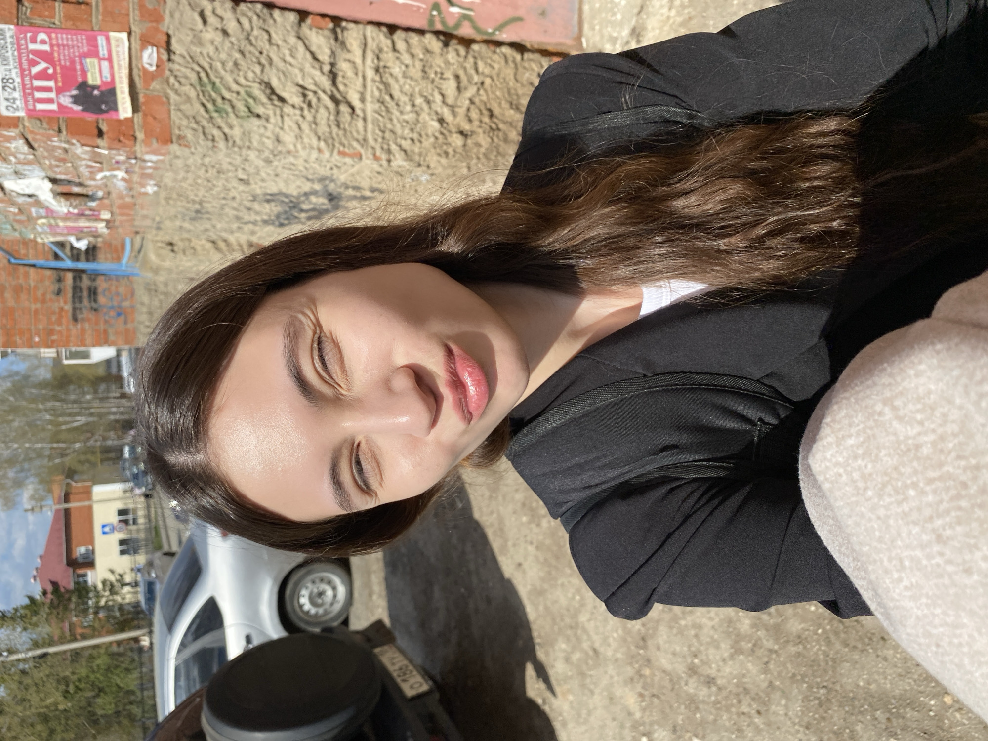 A person, appearing to be a young woman with long, dark brown hair, is the central subject of the image. She is wearing a black jacket and a white shirt, and part of what seems to be a light-colored knitted garment is visible on the right. She is positioned outdoors, with a street scene in the background.
The setting includes a white car, a street with buildings, and trees in the distance, suggesting an urban or suburban environment. A sign in Russian is attached to a brick building, likely an advertisement.
Based on image evidence and available Russian geolocation data, it is possible this image was taken in the city of Votkinsk, Russia. The presence of Russian signage and the architectural style of buildings in the background support this. The scene is captured in bright daylight, casting distinct shadows on the ground. A motorcycle is nearby, casting shadows in front.