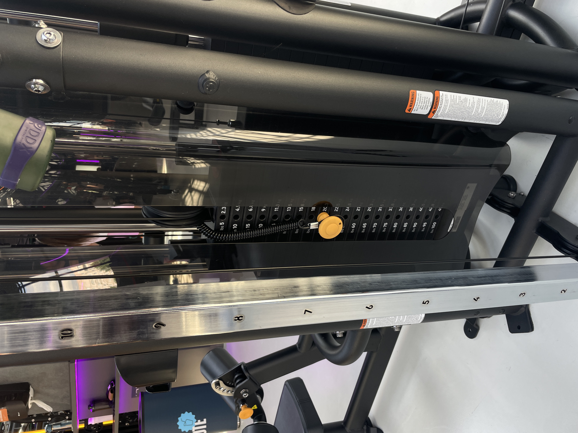The image shows a close-up of a weight machine, specifically focusing on the weight stack and selection mechanism. The weights are stacked in a clear enclosure, with labels indicating weight in both pounds (lbs) and kilograms (kg). A yellow selector pin is inserted into one of the holes, presumably to select the desired weight for the exercise. The machine has a metal frame with a silver rail featuring numbered weight selection points. A warning sticker is prominently displayed on the black frame. The environment appears to be a gym setting, based on the presence of exercise equipment. The location and the city of Kazan, Russia, is not determinable from the picture.