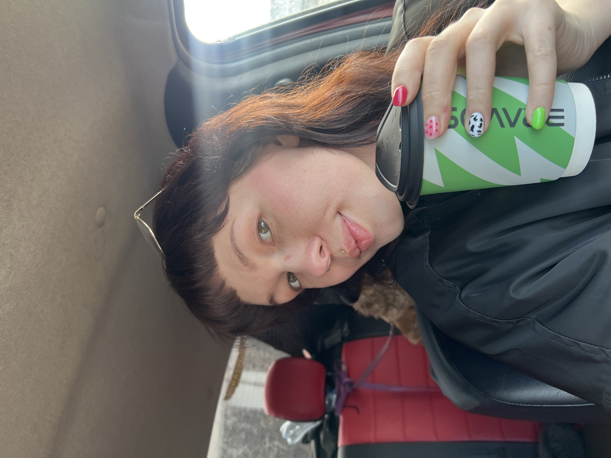 The image shows a person sitting inside a vehicle, possibly a car or van. The vehicle's interior is visible, including the upholstery and a portion of the door. The person is holding a green and white paper cup with a logo that includes the name "BRAVVE" (likely a brand name). They are wearing a black jacket and have distinct nail polish: one nail is red, another has pink dots, one is painted with a black and white cow print pattern, and the fourth is green. The person's face is visible, including lip and nose piercings.
The location is likely Chaikovsky, Russia, based on the presence of the "BRAVVE" logo on the cup.