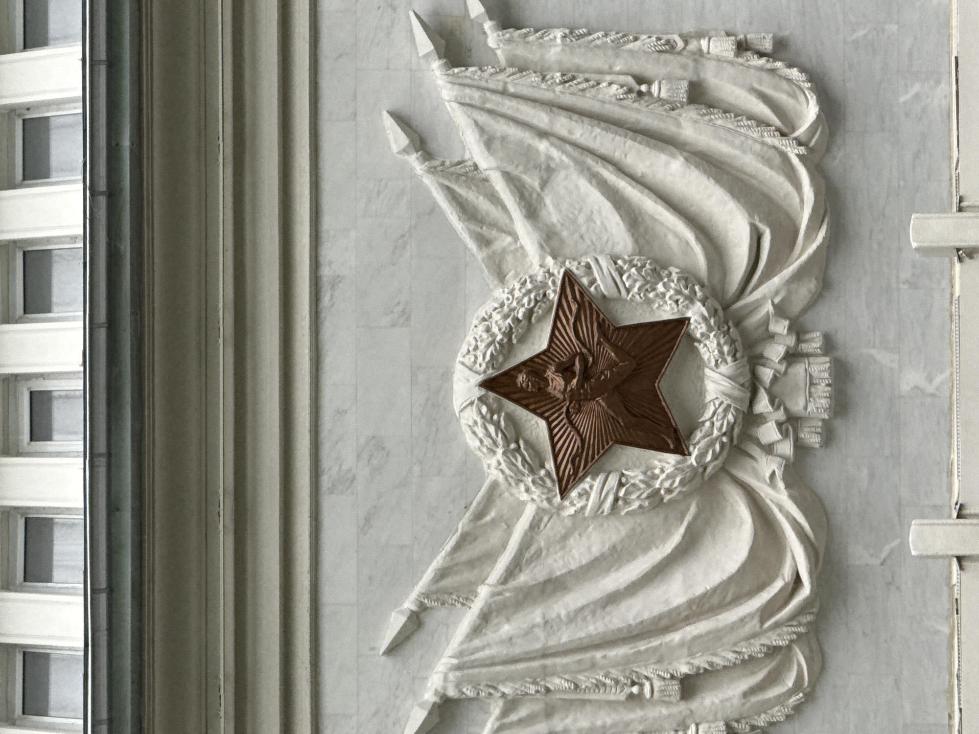 This image shows a close-up of a bas-relief sculpture on a marble building facade. The sculpture depicts a large, bronze, five-pointed star at its center, enclosed by a laurel wreath. Two draped banners, intricately carved with fringe and tassels, flank the star and wreath, extending outwards and downwards. The banners appear to be made of white stone. The architectural detail above the sculpture includes a row of identical windows with dark panes, set into a light-colored, possibly stone, facade. Below the sculpture, more marble blocks are visible, with a hint of a white vertical pillar in the foreground. The overall impression is one of grandeur and monumentality, likely representing a historical or nationalistic theme. Based on the style of the architecture and the prominent Soviet-era star motif, this sculpture is likely located on a building in Moscow, Russia, possibly related to a government or cultural institution from the Soviet period.