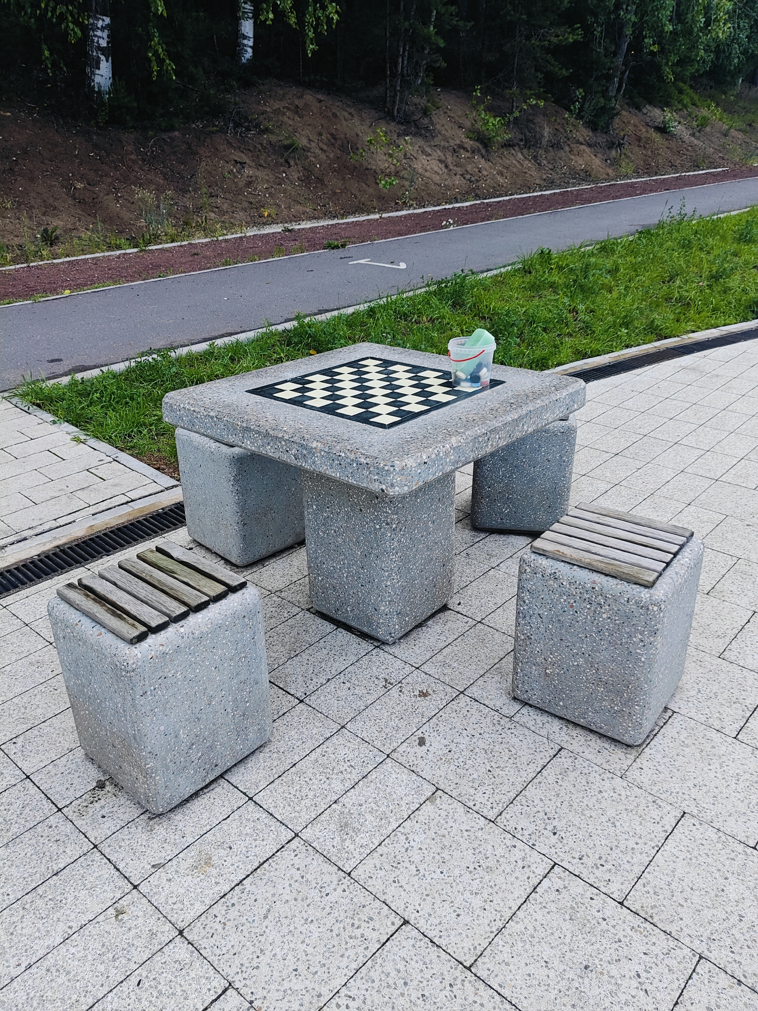 This media file depicts an outdoor chess table with accompanying benches made of speckled concrete. The chessboard surface is inlaid with black and white squares, and a clear plastic bucket containing various items, possibly chess pieces and cleaning supplies, sits on the table. Two concrete cube-shaped benches, each topped with wooden slats, are positioned around the table. The setting appears to be a park or recreational area, with a paved walkway and a grassy area visible in the background. Beyond the grass, there is a dirt embankment with trees and foliage, suggesting a wooded environment. The time of day appears to be daytime, and the weather seems overcast or slightly hazy, as there are no strong shadows. The scene conveys a sense of leisure and potential activity, inviting visitors to play a game of chess. There is no visible text in the image. The location context is Chaikovsky, Russia.