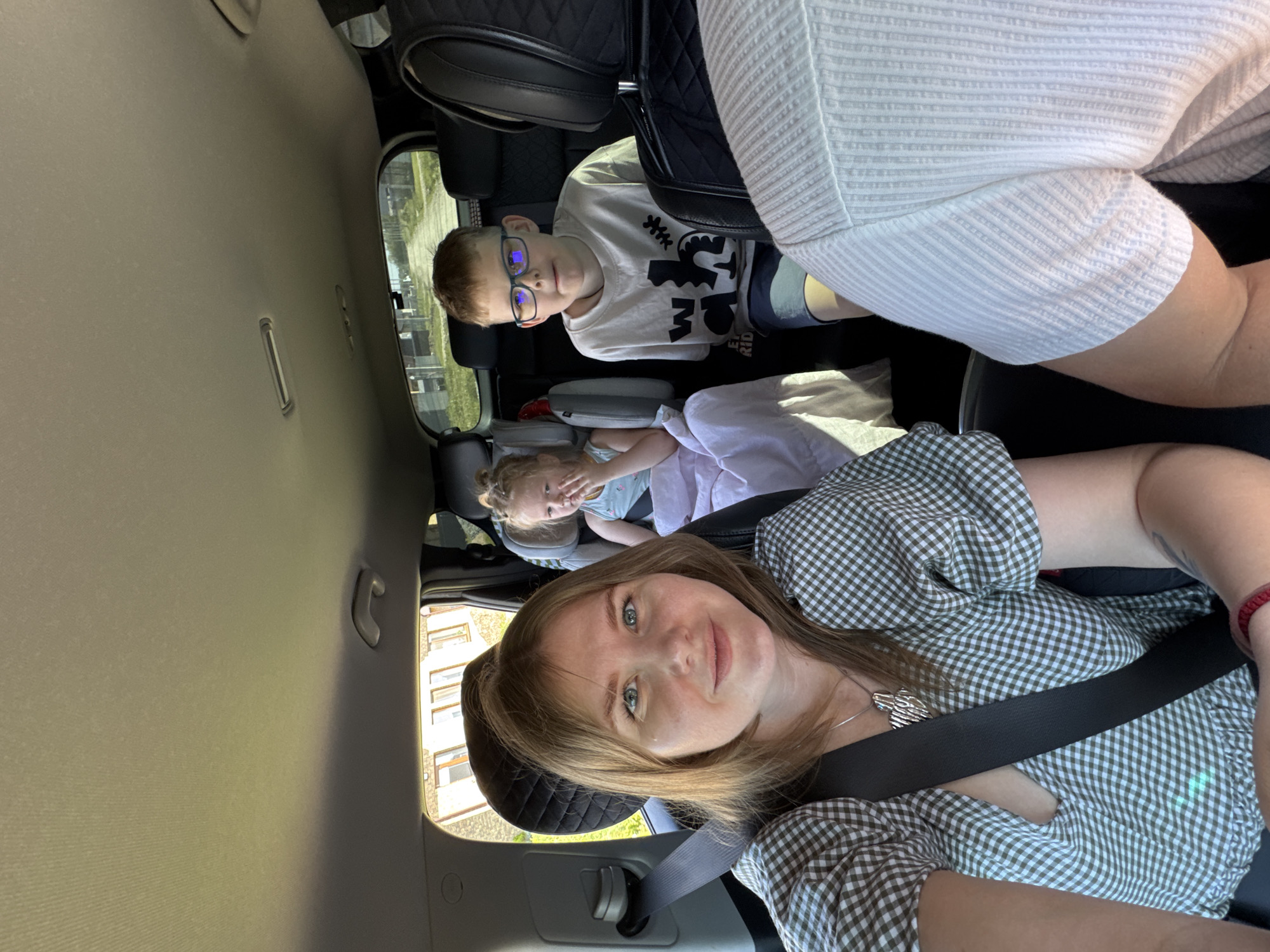 The image shows the interior of a car, seemingly in transit, with a woman in the foreground and two children in the back. The woman in the driver's seat is wearing a green and white checkered dress, a silver necklace, and a seatbelt. She appears to be taking a selfie. In the back, a boy with glasses is seated behind the driver's seat, and a younger girl lies down behind the passenger seat. The location is inferred from previous context to be near Горный, Russia, based on the presence of buildings visible through the car windows, but is not directly visible.