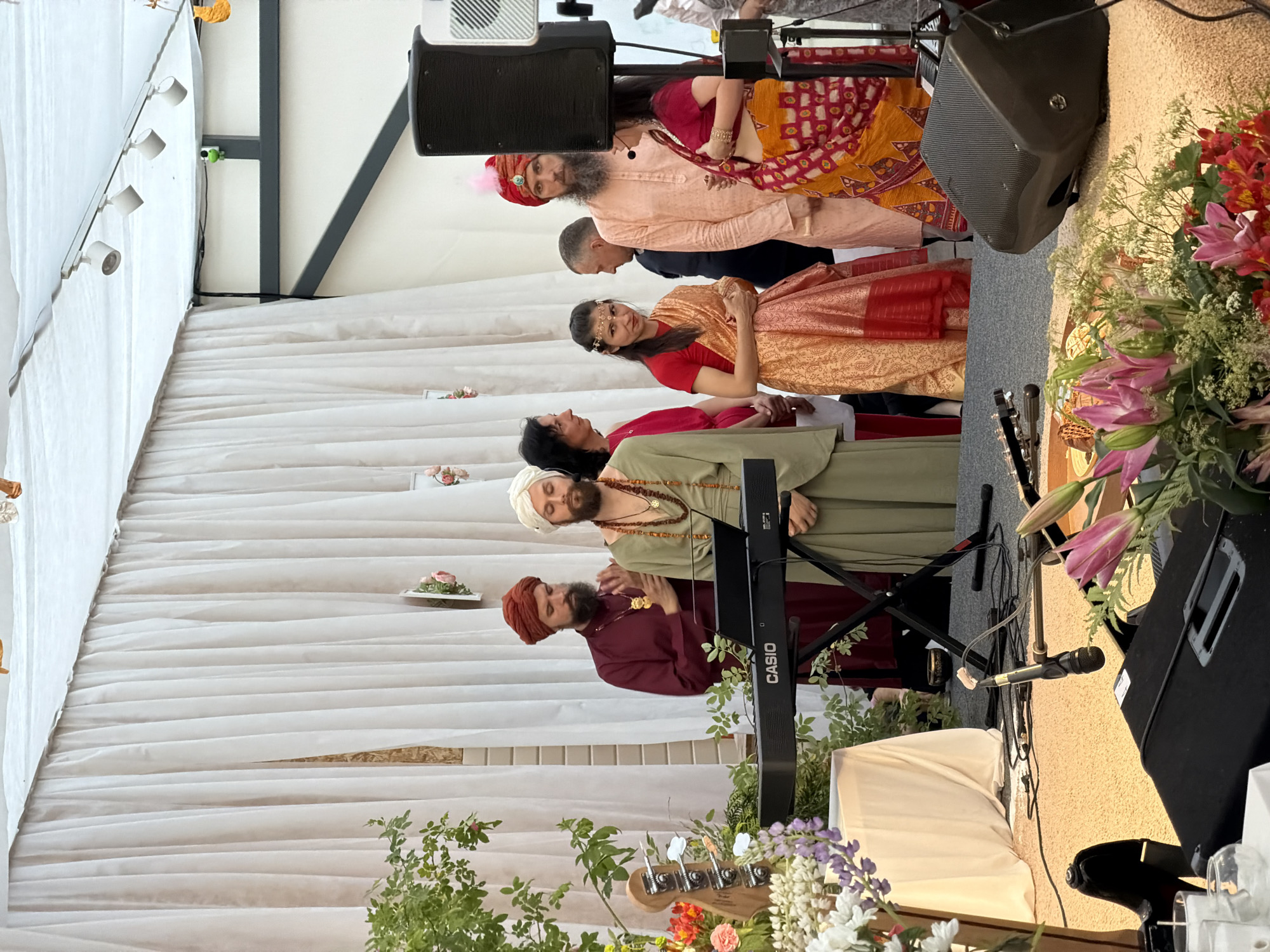 In the city of Zhirnosovo, Russia, a group of individuals are gathered outdoors. White drapes hang in the background, suggesting a temporary structure or tent. In the foreground, a keyboard is set up on a stand, with a microphone attached to an arm. A bass guitar lies on the ground near a speaker. Several people are visible, some dressed in traditional Indian attire, including a man in a maroon robe and turban, another in a peach-colored outfit and pink turban, and a woman in a red and gold sari. Another man, wearing a light green robe and a white turban, stands behind the keyboard. The scene appears to be a performance or a cultural event, possibly a wedding or religious ceremony, with floral arrangements adding to the festive atmosphere.