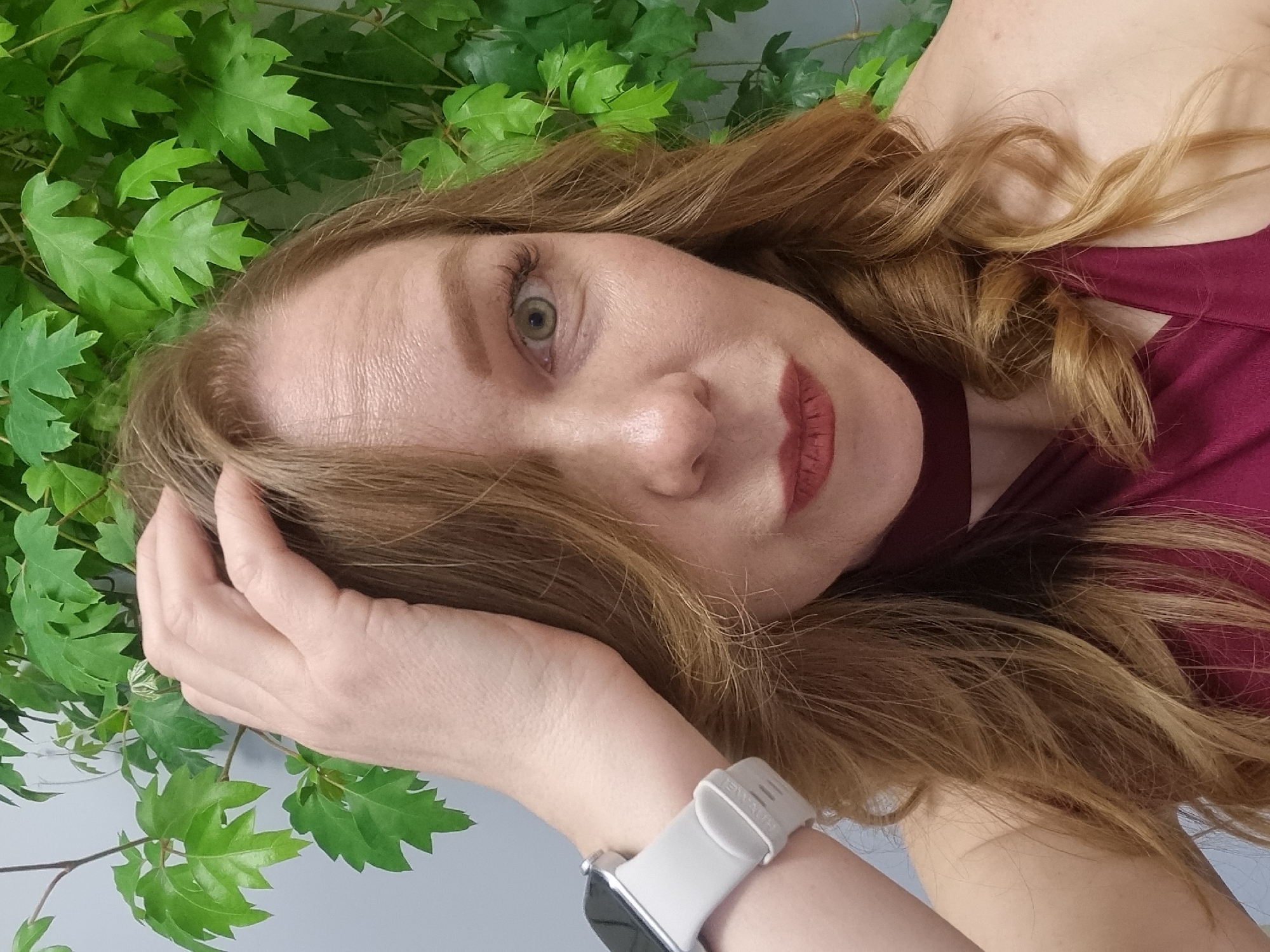 A light-skinned woman with reddish-brown hair and light-green eyes is posing in front of a wall covered in green, leafy vines. She is wearing a maroon top and a white smartwatch on her left wrist. Her left hand is touching her forehead. The image is taken from an oblique angle and appears to be a close-up portrait. The city of Kemul, Russia, is not present in the image.