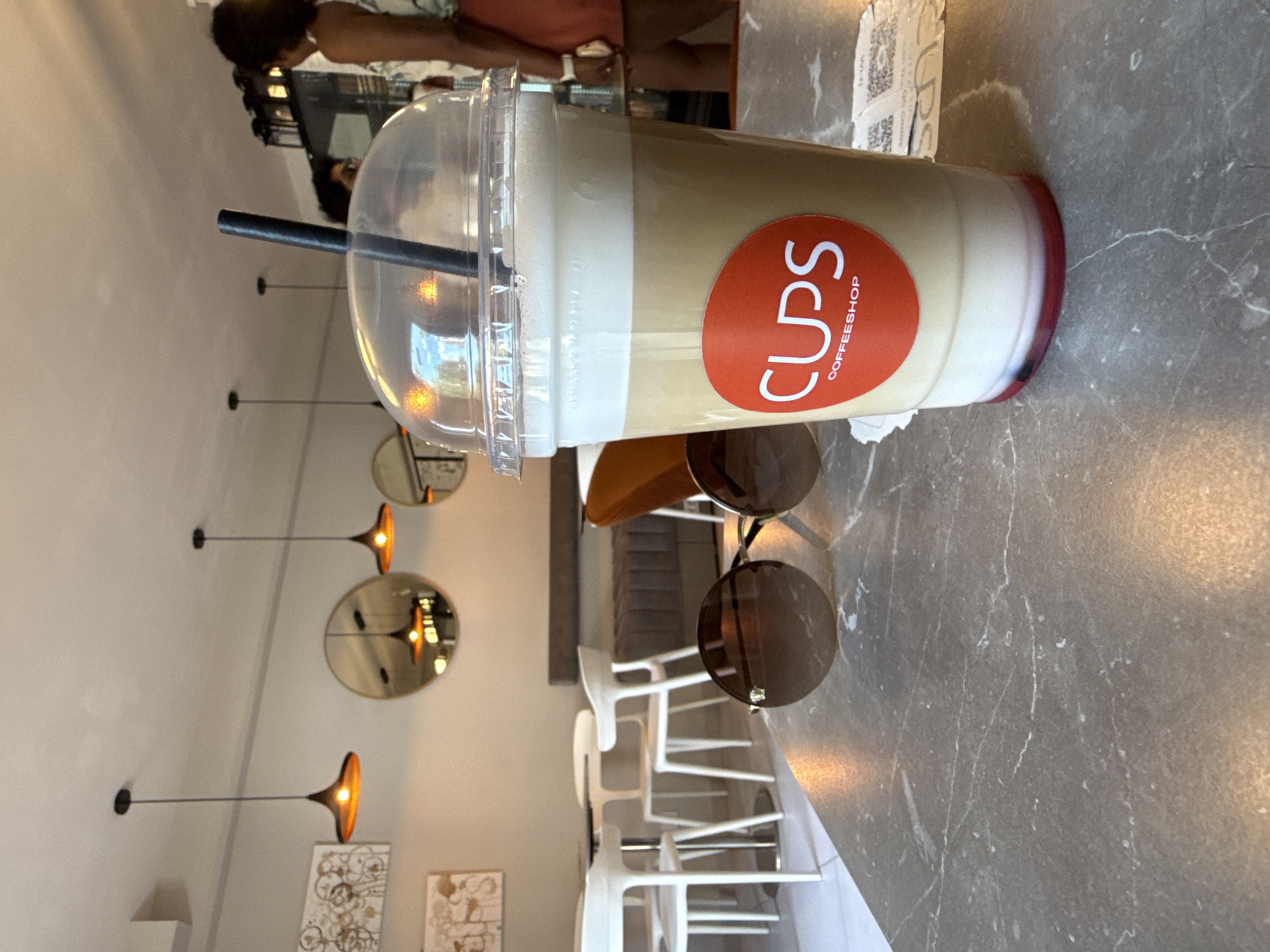 This media file depicts a close-up view of a clear plastic cup filled with a layered beverage, likely a coffee drink with foam, on a marble countertop. The cup has a red circular logo with white text that reads "CUPS COFFEE SHOP". A dark straw is inserted into the cup.
In the background, hints of a cafe setting are visible. There are several modern white chairs arranged around tables, suggesting a place of business. Hanging pendant lights with warm illumination are also present, casting a soft glow. Two circular mirrors with reflective surfaces are mounted on a light-colored wall, one reflecting the lights and the other reflecting more of the cafe interior.
The overall scene appears to be indoors, within a coffee shop. The lighting suggests it could be daytime or evening, as the interior lights are on. The atmosphere seems relaxed, typical of a cafe environment. While no specific event is evident, the presence of the beverage and the seating arrangement indicate a moment of casual enjoyment or respite. The image is oriented such that the cup is tilted, with the logo prominently displayed.