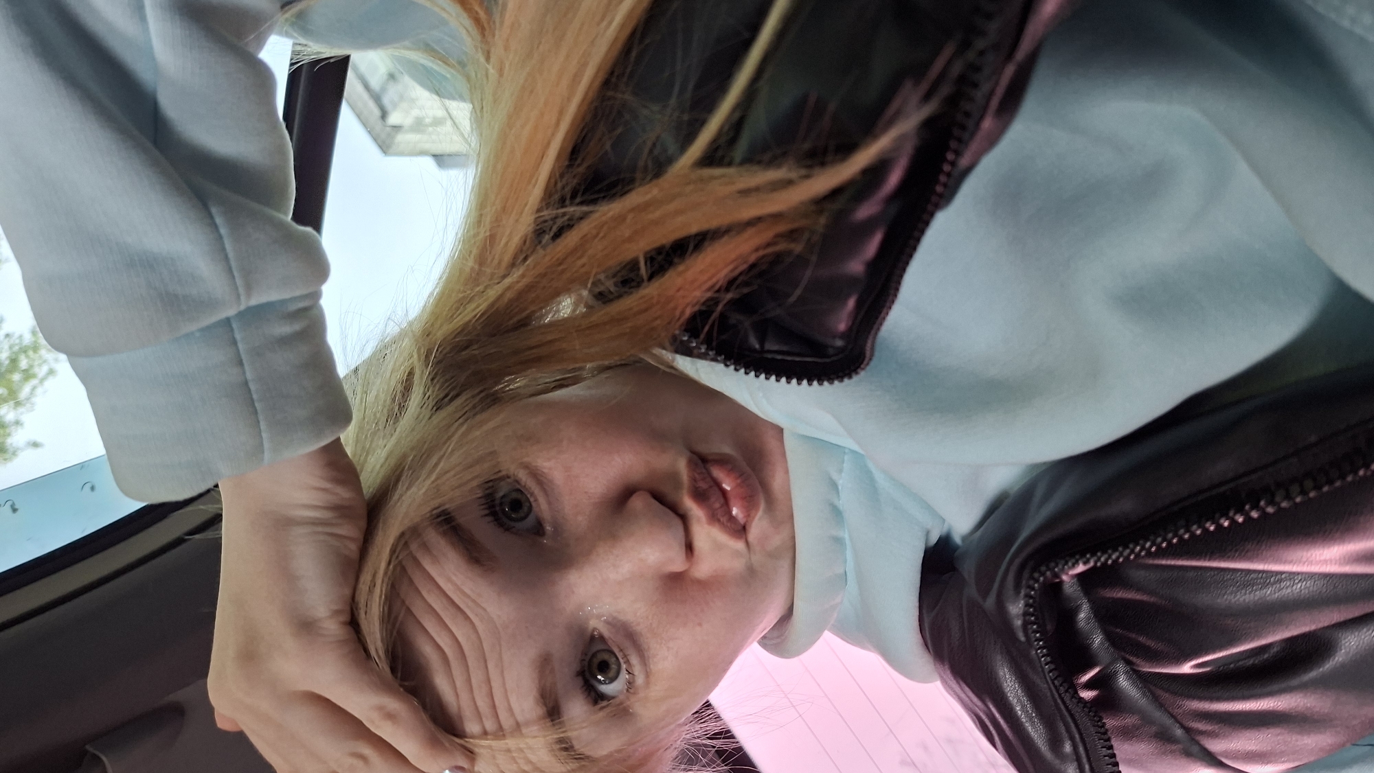 A woman is depicted from a close, tilted, and upside-down perspective. She has fair skin, light-colored hair, and appears to be wearing a light blue hoodie partially covered by a dark, possibly leather jacket. Her face is prominent, showing details such as her eyes, nose, and mouth. Part of a car's interior is visible in the background, specifically the window and roof. It is not possible to determine the location as Chaikovsky, Russia, from visual evidence alone.